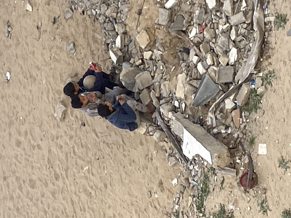 The image displays an outdoor scene in Khan Yunis, Palestinian Territory, featuring three young males situated on a mound of debris. The ground consists primarily of light brown sand, sparsely littered with small pieces of trash and rocks. The subjects are positioned on an elevated pile of concrete rubble, broken stones, and twisted metal fragments.
The leftmost individual wears a dark blue hooded jacket and light grey pants, crouching with his hands near a small object in front of him. The central individual wears a grey and white striped long-sleeved shirt and grey pants, crouching and holding his hands near his face, looking towards the right. The rightmost individual wears a dark blue jacket and pants, crouching while holding a light-colored spherical object under his left arm and extending his right hand, which holds a small red object, towards the ground or the other children.
The rubble mound includes large, irregularly shaped concrete pieces, smaller stone fragments, and bent metal elements. In the lower left foreground, a red, rusted, wheel-like object is visible amidst the sand. Scattered pieces of litter, including paper and plastic, are present across the sandy ground and within the debris. Sparse green vegetation is visible emerging from the sand in the lower sections of the frame.