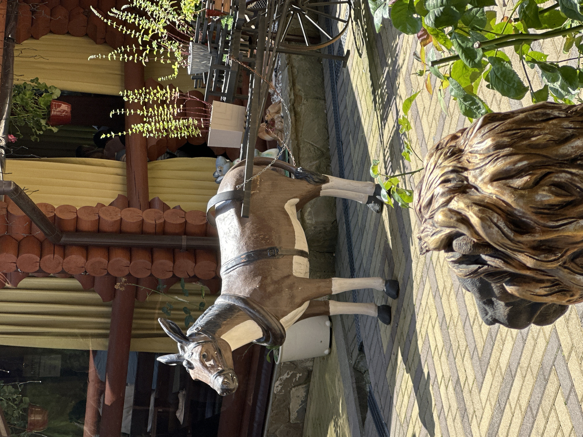 The image depicts a scene outside a log cabin structure in Krasnaya Polyana, Russia. In the foreground, a statue of a lion's head is partially visible. Behind it, a life-sized statue of a brown and white donkey stands tethered to a dark metal cart. The donkey statue is equipped with a harness and bridle. The cart has large spoked wheels. The ground is paved with interlocking brick tiles. In the background, yellow curtains hang in a window of the log cabin, and several potted plants are arranged on a ledge. The overall impression is that of a decorative outdoor setting, possibly at a cafe or resort.