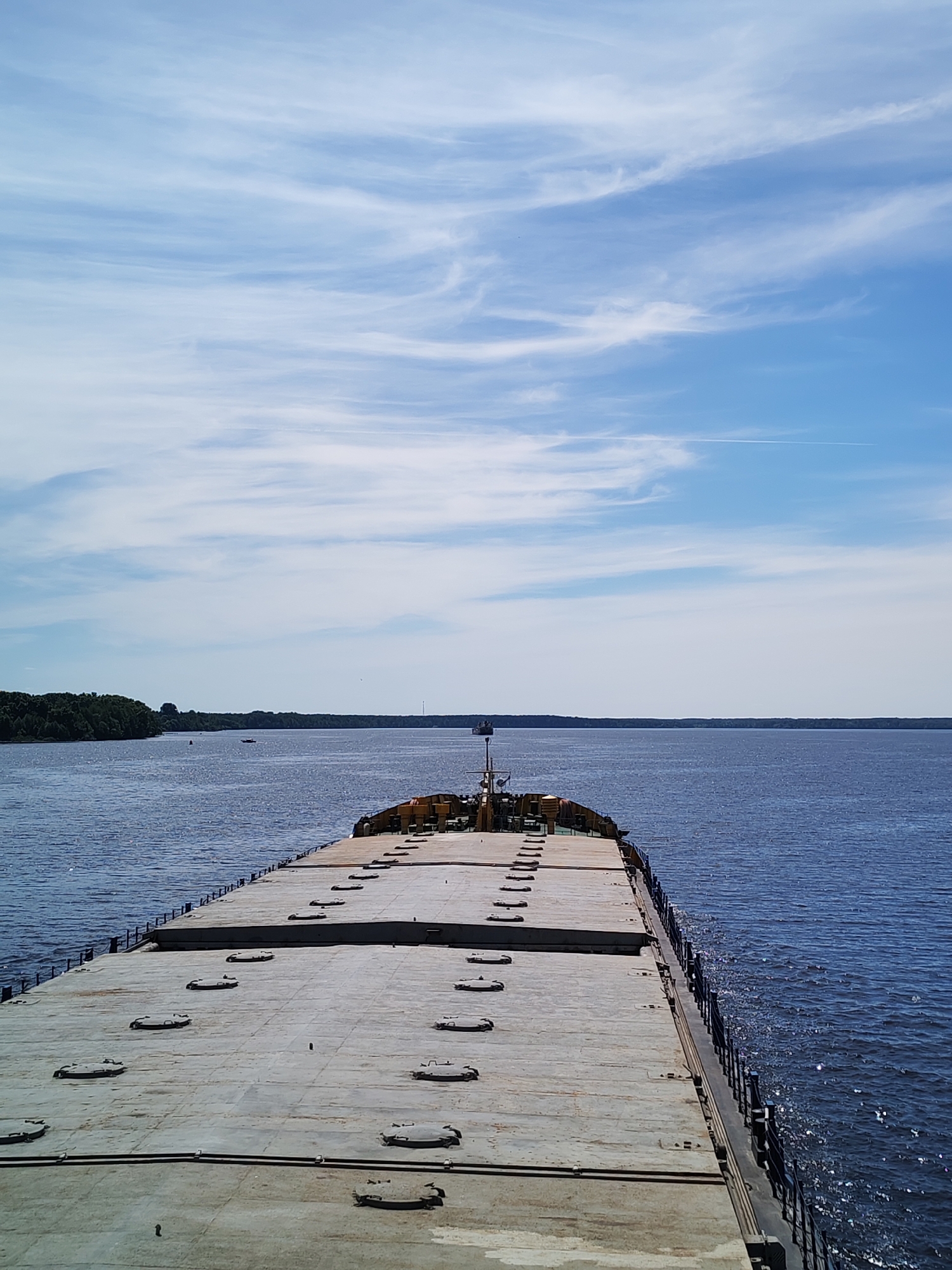 The image depicts the forward deck of a large, flat-topped cargo vessel, likely a barge or bulk carrier, underway on a wide body of water. The deck, a light grey concrete or metal surface, features numerous evenly spaced, oval-shaped hatch covers. A series of dark vertical stanchions line the port side of the deck, forming a simple railing, with a similar structure visible on the starboard side. Towards the far end of the vessel, a small superstructure or pilot house is visible, with yellow and green elements.
The vessel navigates a vast, calm expanse of dark blue water, reflecting scattered sunlight. Distant landmasses, heavily forested with green trees, line both the port and starboard horizons, suggesting a wide river or a large lake. A small red boat and another dark, indistinct vessel are visible in the distance on the port side, closer to the tree line. On the starboard side, further down the waterway, a dark, vertical structure, possibly a mast or tower, rises above the distant tree line.
The sky above is a clear, light blue with delicate, wispy white cirrus clouds stretching across the frame, indicating fair weather. A faint, linear contrail from an aircraft is visible high in the sky towards the upper right. The scene is set in the city of Priluki, Russia, on this expansive waterway.