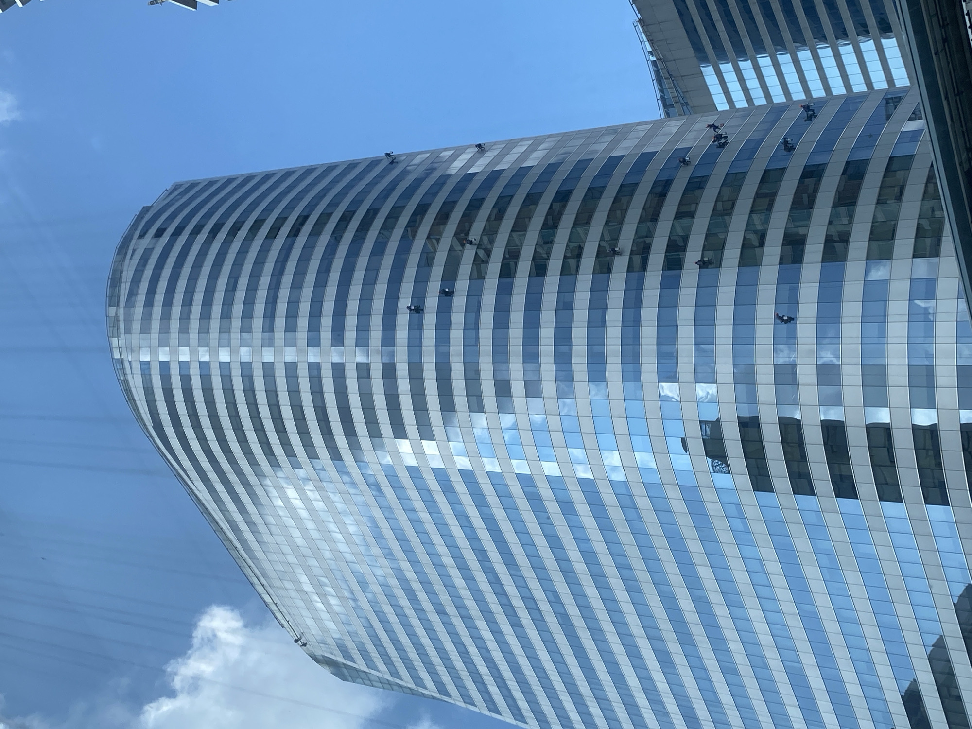 This is a low-angle shot of the exterior of a curved skyscraper in Bangkok, Thailand. The building has a glass facade with vertical white stripes that create a textured appearance. There are several window washers on the building, suspended from ropes. The sky is blue with a few white clouds. Another skyscraper can be seen at the top of the frame.
