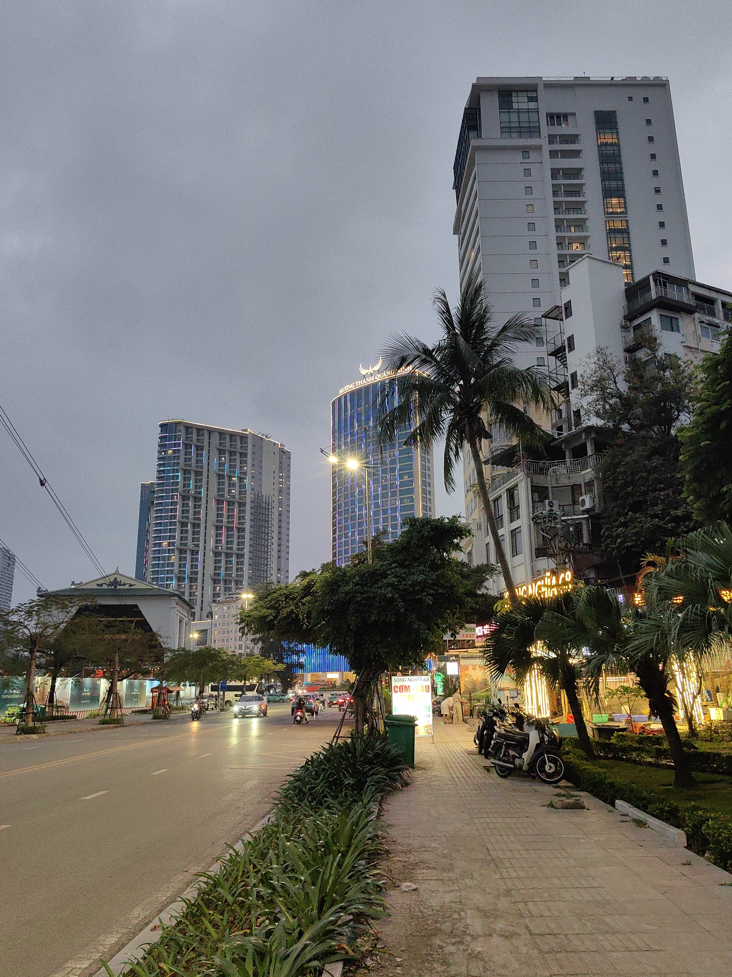 The image depicts a street in Bai Chay Ward, Vietnam at dusk. A multi-lane road runs alongside a sidewalk with vegetation and a row of palm trees. The road is mostly empty, with a few cars and motorbikes driving on it. The sidewalk is lined with plants and trees. Several motorbikes are parked on the sidewalk near businesses with illuminated signage. Tall buildings loom in the background, including the Muong Thanh hotel. The sky is overcast.