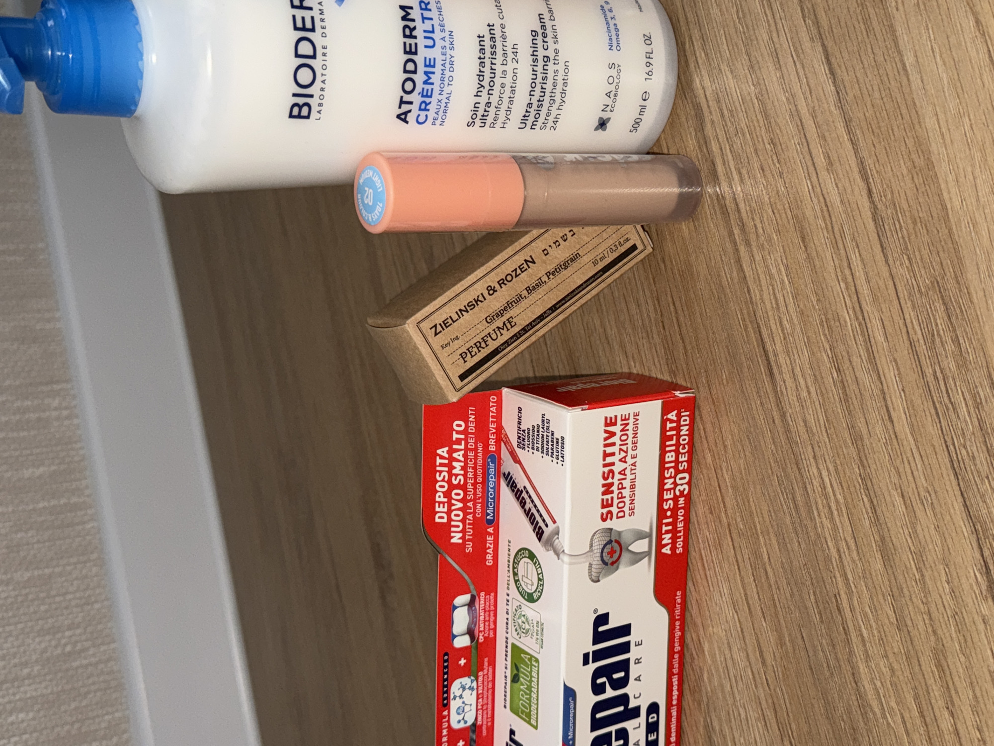 The image shows a close-up view of several cosmetic and personal care products arranged on a wooden surface. In the foreground, a tube of "Biorepair" toothpaste is positioned horizontally, with its packaging prominently displaying information about enamel repair and sensitive teeth. To the right of the toothpaste, a small cylindrical bottle with a peachy-pink cap stands upright. Behind these items, a large pump bottle of "Bioderma Atoderm Creme Ultra" moisturizer is visible. To the left of the peachy-pink bottle, a small, rectangular brown cardboard box, labeled "ZIELINSKI & ROZEN PERFUME," is propped up. The background consists of a light-colored textured surface, possibly a wall or a piece of furniture, with a subtle horizontal line detail. The lighting is soft, casting gentle shadows on the wooden surface. There are no people or animals visible in the image. The image appears to be taken indoors, with no indication of a specific location in the city of Perm, Russia.