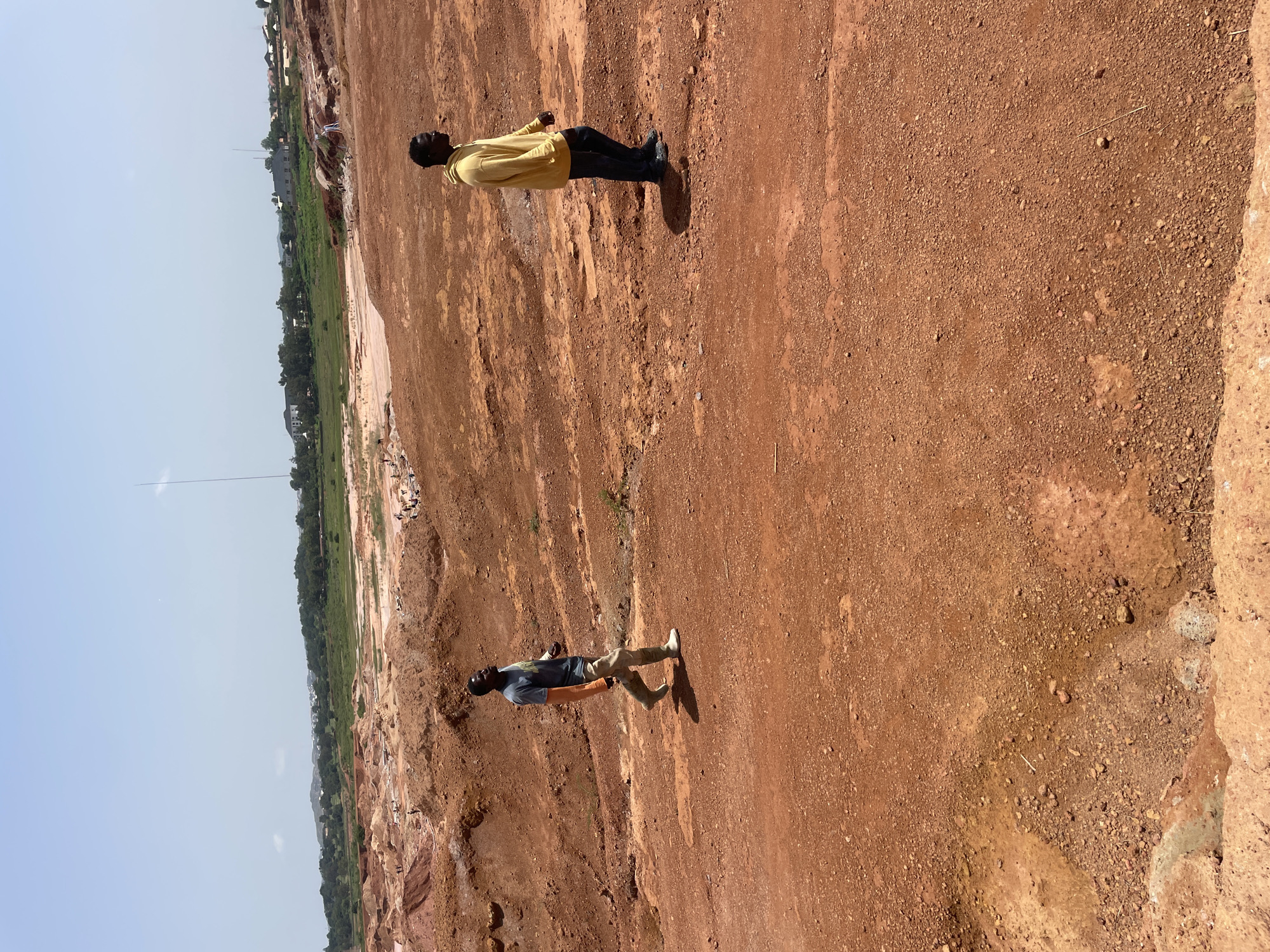 The image shows two men standing in a dusty, outdoor environment. The ground is composed of red earth and small stones, characteristic of a construction site or a natural, arid landscape. In the background, there's a view of a valley with green vegetation, some buildings, and a tall, thin structure that could be a communication tower or a flag pole. The sky is clear and bright, suggesting a sunny day. The overall scene depicts an open, undeveloped area. The men appear to be observing the surroundings or perhaps engaged in a conversation. One man is wearing a yellow long-sleeved shirt and dark pants, while the other is in a blue t-shirt and light-colored pants. The image appears to be taken during daylight hours. There is no visible text in the image. The location context is Jos, Nigeria.