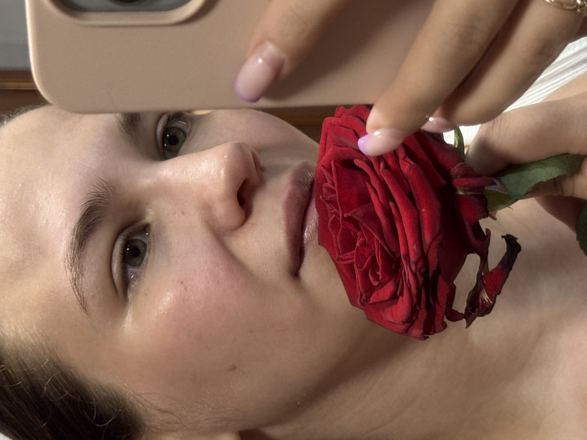A close-up, frontal selfie shows a young woman holding a red rose and a light pink smartphone. She is looking at the phone's camera, which is off to the right of the frame. The woman has fair skin, light brown hair pulled back, and green eyes. She is wearing a white top. The rose is held near her mouth, with its petals extending towards the bottom left of the frame. Her right hand is visible, holding the phone, and her left hand is supporting the rose. The background is blurred and appears to be an indoor setting, possibly a bedroom, in Chaikovsky, Russia.