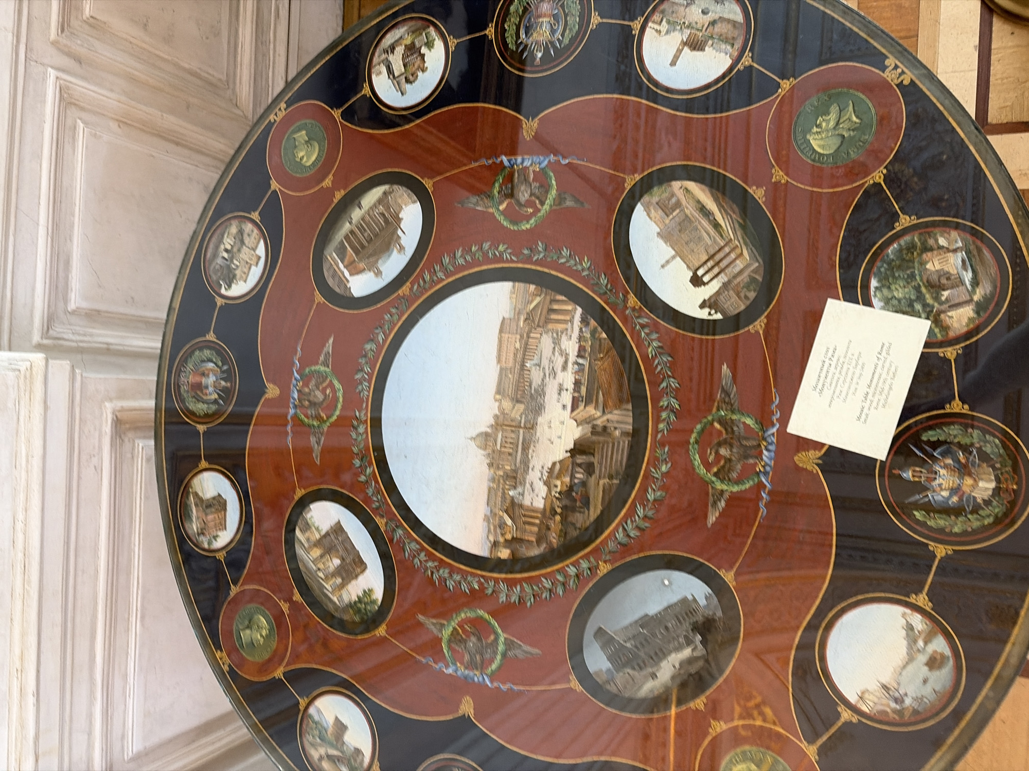 The image depicts a round mosaic table. The table is richly decorated with images of Roman monuments in the central part and along the perimeter. The color palette includes red, black, and gold. There's a white card with printed text lying on a section of the table near one edge. The table appears to be located indoors, possibly in a museum or historical building, as indicated by the adjacent white wall with detailed panels.