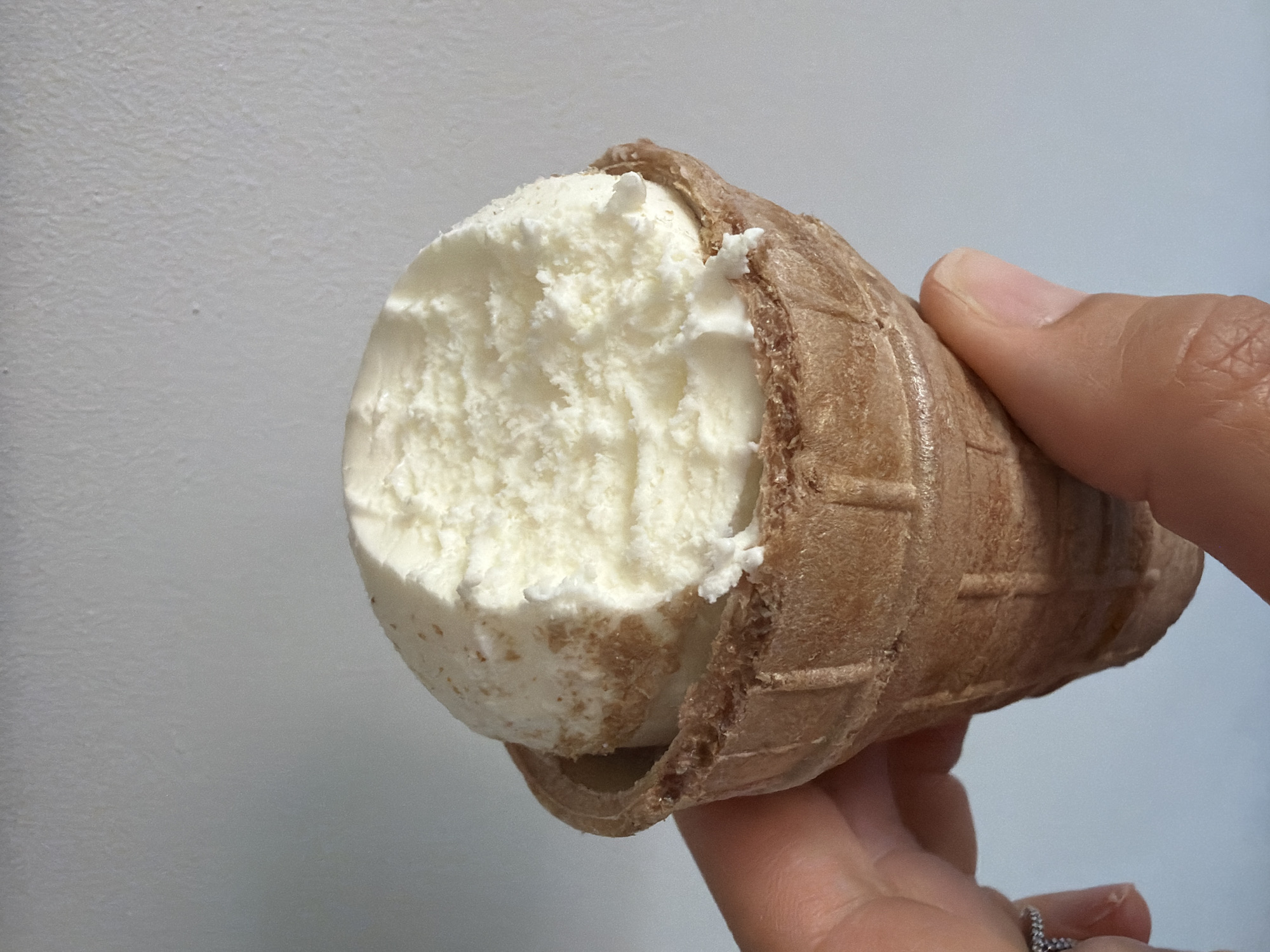 A close-up shot shows a hand holding an ice cream cone filled with vanilla ice cream against a plain, light-colored wall. The ice cream has a bite taken out of it. The waffle cone is a light brown color and has a textured surface. The hand is visible from the wrist down and wears a ring on the index finger. The background wall appears to have a slight texture. This image does not provide any information about the location in the city of Chaikovsky, Russia.
