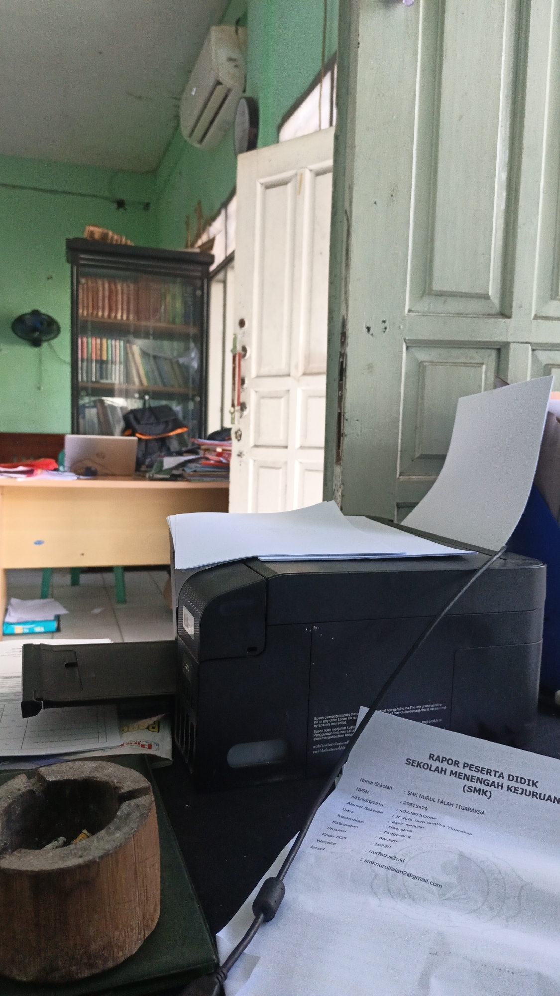 The image depicts an indoor scene that appears to be an office or classroom in Tigaraksa, Indonesia. In the foreground, a black printer is placed on a desk, with a sheet of paper extending upwards from its output tray. To the left of the printer, an ashtray made of wood is visible. The desk also holds a document with printed text, including "SMK NURUL FALAH TIGARAKSA", indicating the location. In the background, a wooden desk with a laptop, books on shelves in a cabinet, and a wall-mounted air conditioner are discernible. The overall environment suggests a workspace that may also serve as an educational setting.