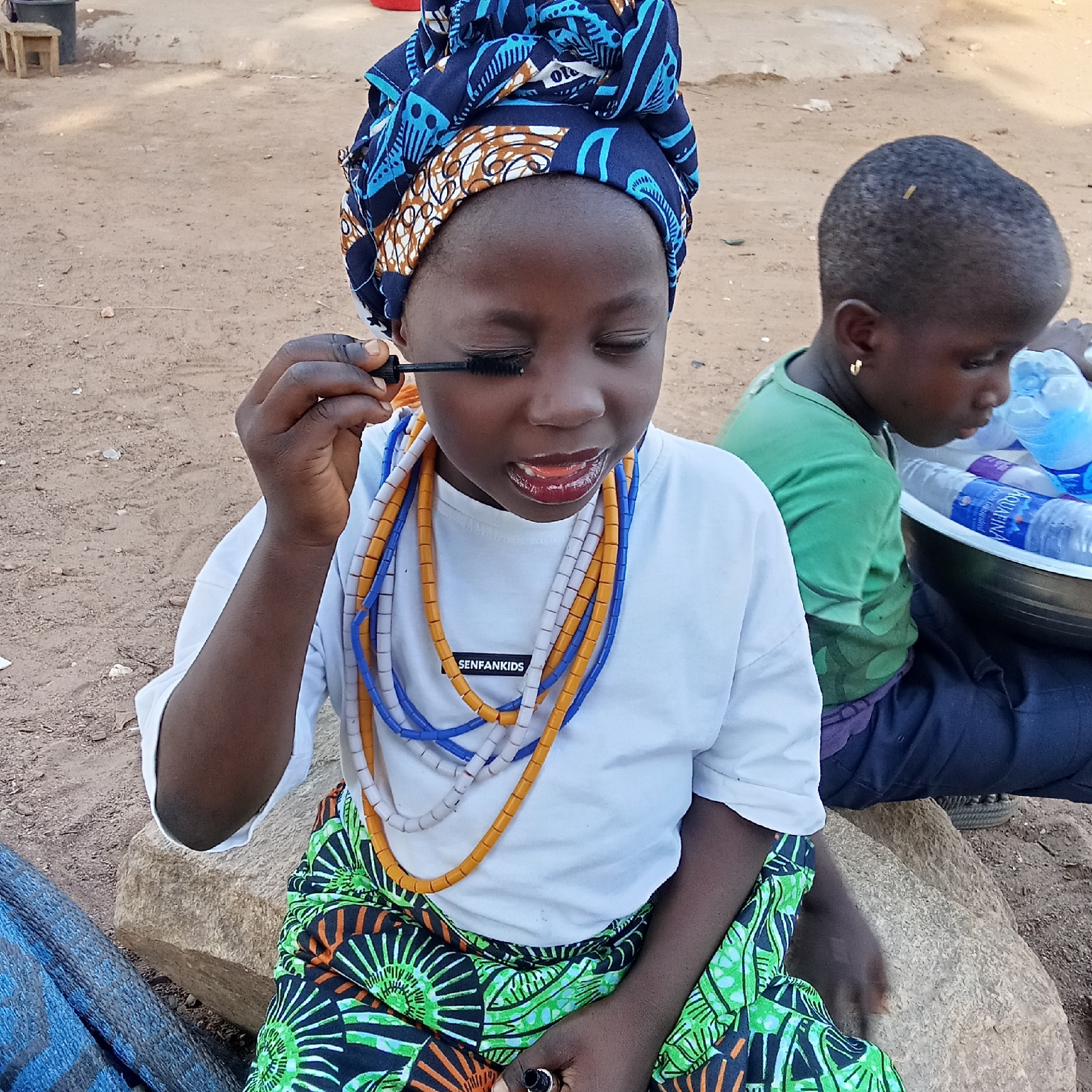 In an outdoor setting in Akwanga, Nigeria, a young girl with dark skin is prominently featured in the foreground, focused on applying mascara to her eyelashes. She is wearing a white t-shirt with "SENFANKIDS" printed on it, a colorful green, black, and orange patterned skirt, and a distinctive blue and brown patterned headwrap. Several strands of beaded necklaces in blue, white/grey, and orange adorn her neck. Her eyes are closed in concentration, and her mouth is slightly open, as she holds the mascara wand. She sits on a large, light-colored rock.
In the background, to the right, another young child, also with dark skin and short dark hair, is seated on a similar rock. This child wears a green t-shirt, dark pants, and a small gold hoop earring, and appears to be looking down at a large bowl filled with plastic water bottles, some of which are branded "Aquafina."
The ground is dusty and reddish-brown, indicative of a rural or semi-urban outdoor environment. In the far left background, a black bucket and a small wooden stool are partially visible. The scene is brightly lit, suggesting it is daytime. The primary girl's act of applying makeup, possibly mimicking adults, lends a playful and observant tone to the image.