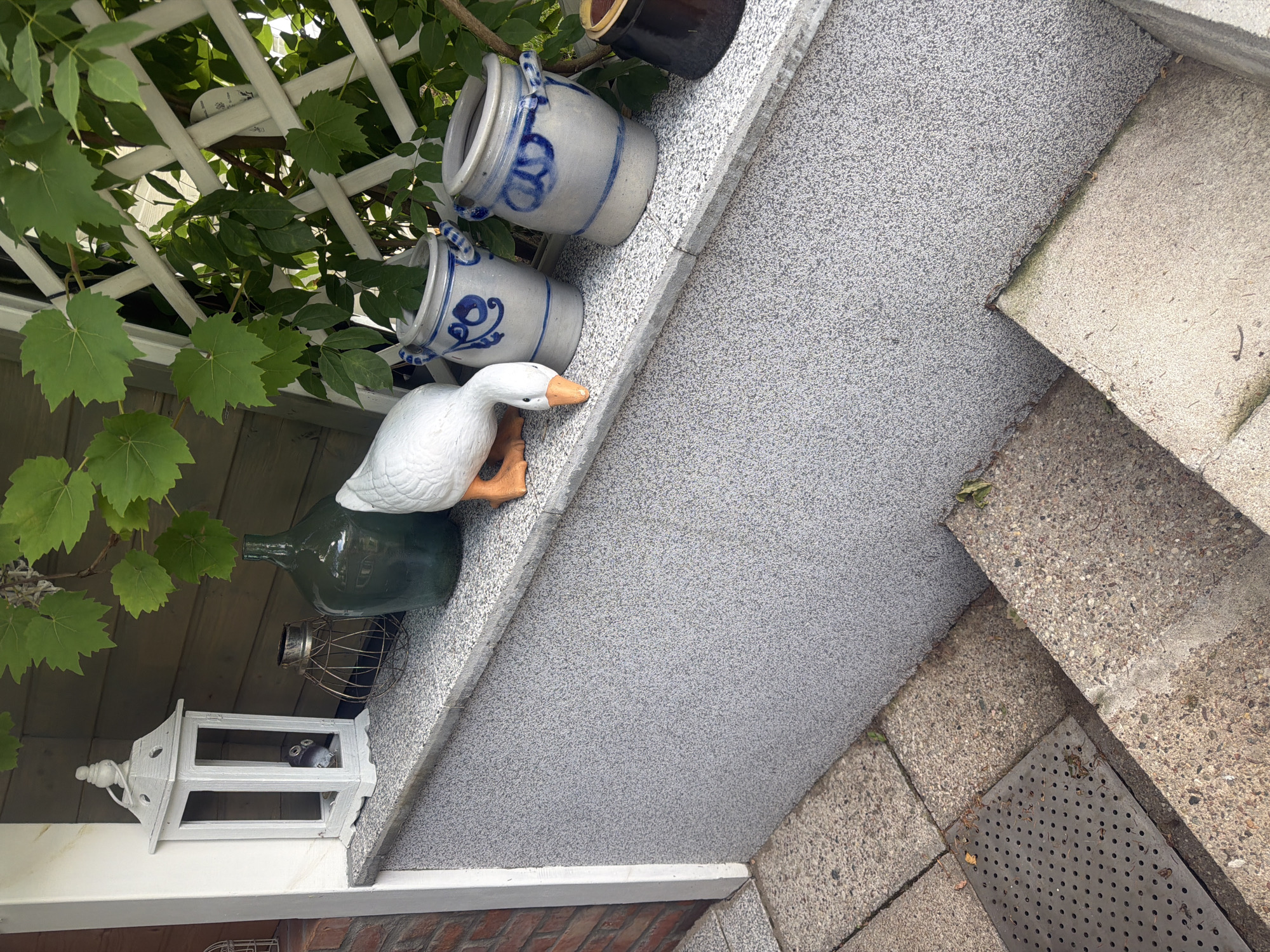 The image depicts an outdoor scene featuring a collection of decorative items on a ledge beside a textured gray wall. On the ledge, a white lantern with a pointed roof is visible to the left, containing a small dark object inside. Next to it is a round wire cage and a large, dark green glass bottle. Further to the right, a white ceramic goose with an orange beak is positioned, with its head down as if grazing. Behind the goose are two stoneware pots with blue decorative patterns. A white lattice structure and green foliage are present in the background. The ledge appears to be a decorative element of a building, with steps leading down to a paved area in the foreground. The overall setting suggests a garden or entryway. The image was taken in Międzyzdroje, Poland, a seaside resort town on the Baltic coast.