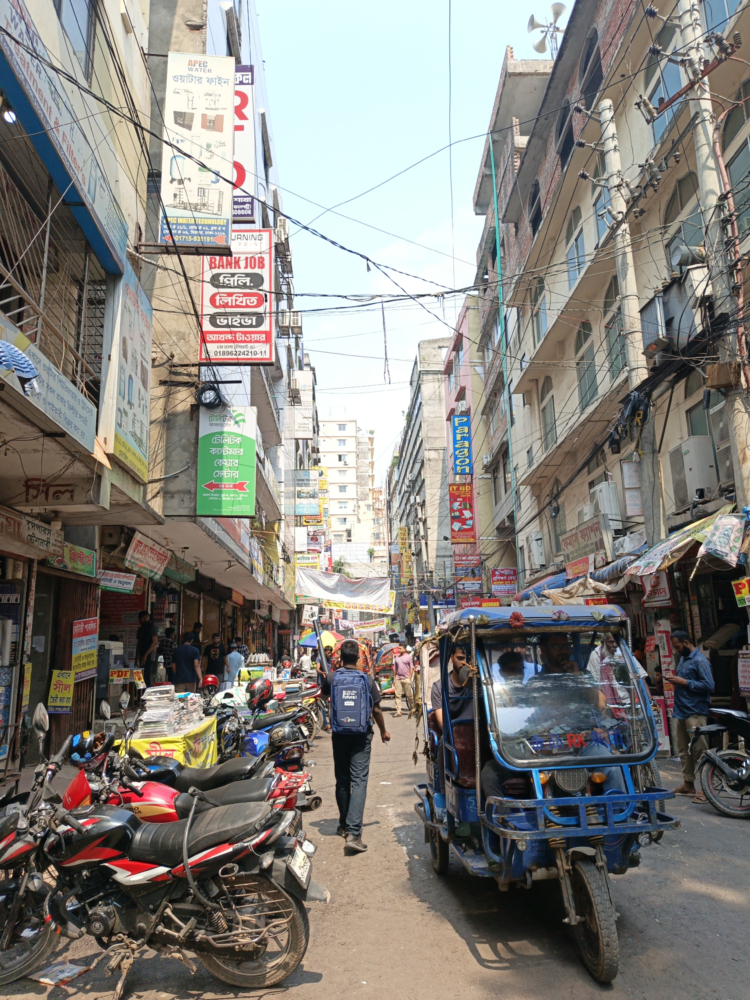 The image depicts a busy street scene in Dhaka, Bangladesh. The narrow street is crowded with pedestrians, motorcycles, and a blue three-wheeled motorized rickshaw, carrying several passengers. The street is lined with shops and businesses, with numerous signs in Bengali advertising various products and services, including water treatment, bank jobs, and a Teletalk customer care center. Overhead, a tangled web of electrical wires crisscrosses between buildings. The buildings are a mix of aged and newer structures, with exposed brick and concrete. The sky is a hazy blue. People are walking in different directions, with one man in the foreground wearing a blue backpack. Several motorcycles are parked alongside the street. The image is well-lit, suggesting a sunny day.