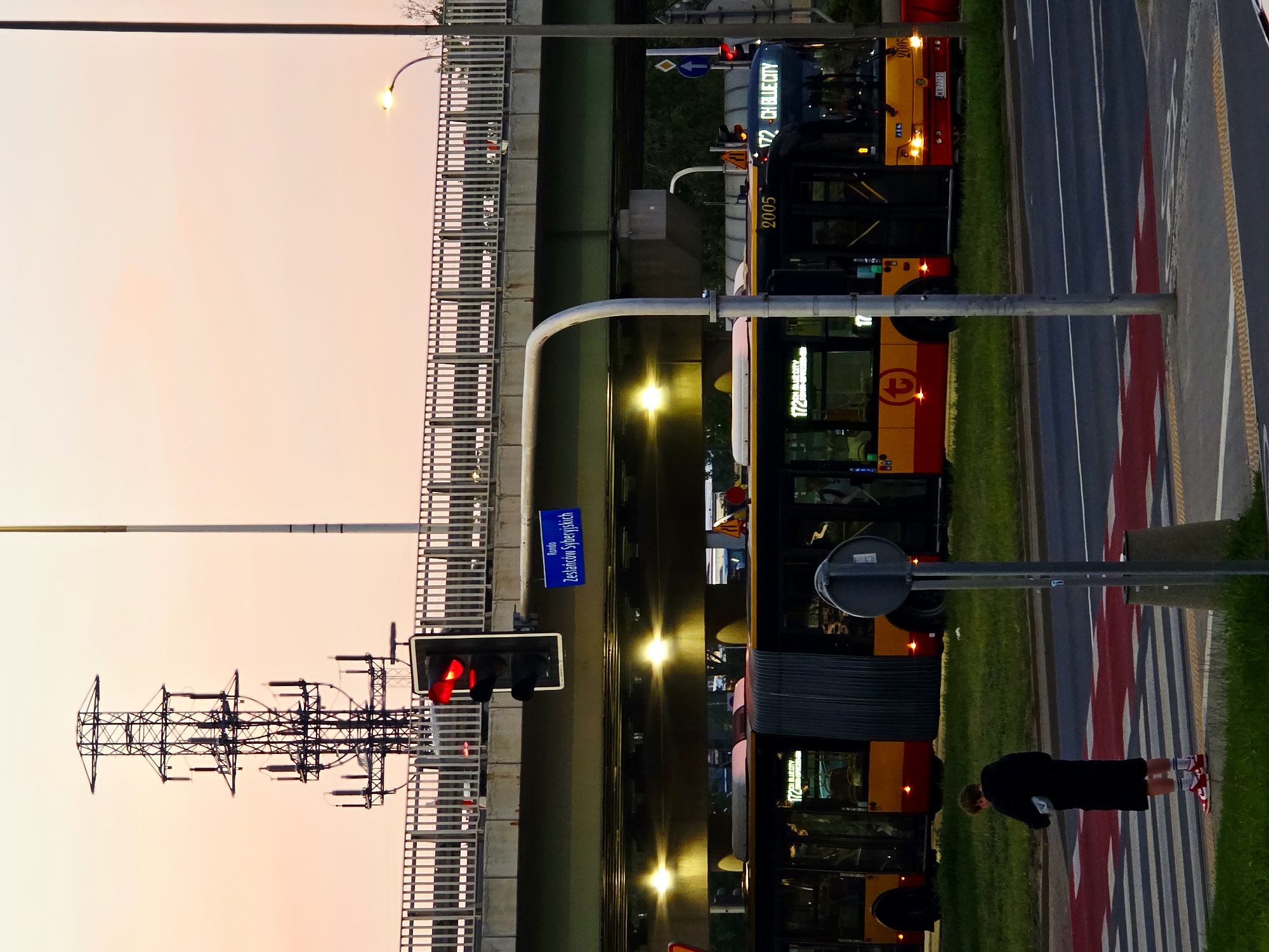 The image shows a city street scene at dusk in Warsaw, Poland. A person stands on a crosswalk. Two buses are stopped at a bus stop under an elevated road. A traffic light is red. A street sign is visible on the elevated road. The sky is a pale pink.