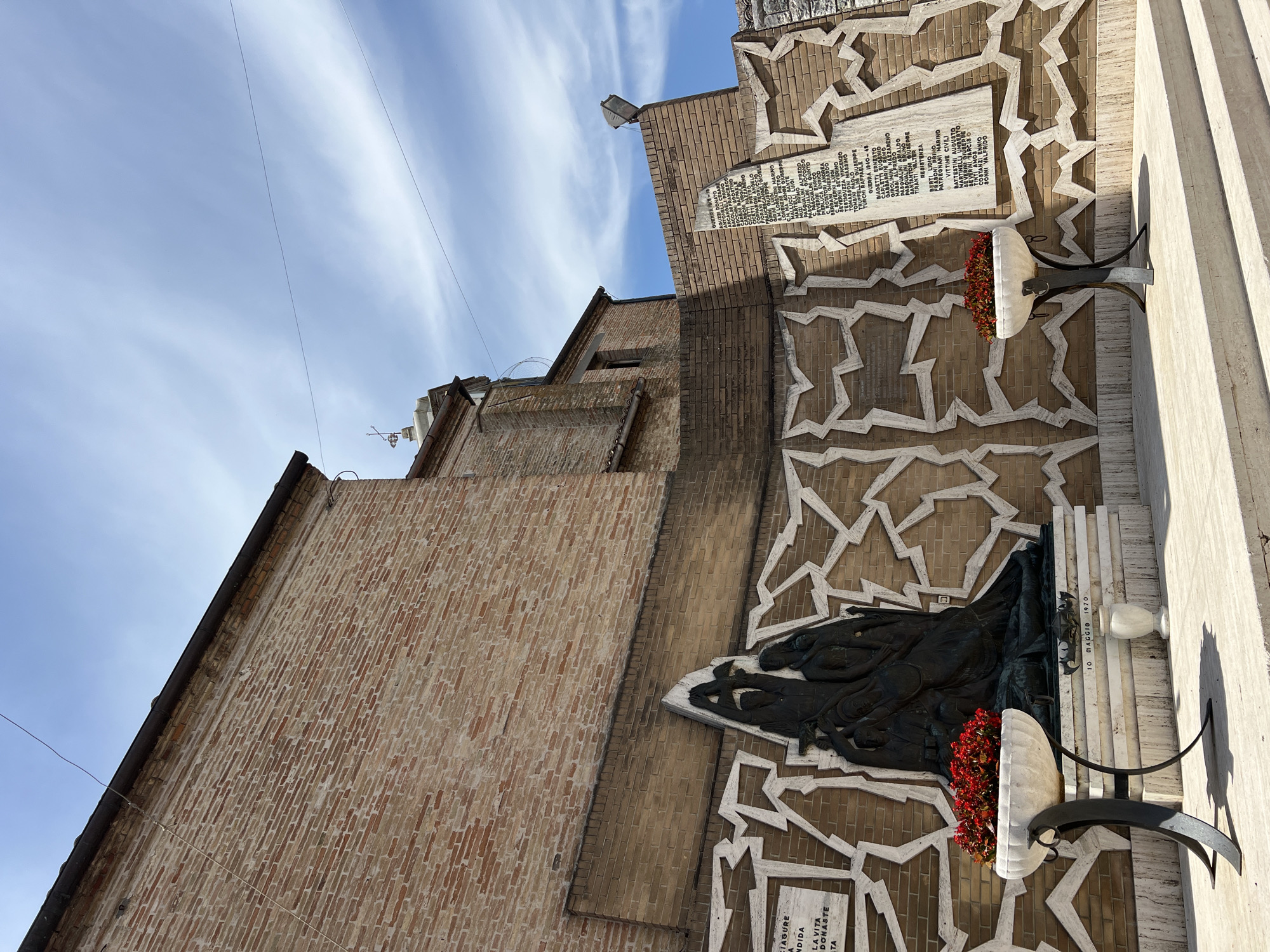 This is a photograph of a war memorial located in the city of Barbara, Italy. The memorial is made of brick and features a bronze sculpture of figures, possibly a family or group of people, in distress. The sculpture is placed on a tiered stone base in front of a brick wall decorated with a white geometric pattern. To the left of the sculpture, a marble plaque is mounted on the wall, bearing an inscription in Italian. Two large white vases filled with red flowers are positioned on either side of the sculpture, serving as planters. To the right of the sculpture, another marble plaque is affixed to the wall, also inscribed with Italian text. The memorial is situated outdoors, with a blue sky and white clouds visible in the background, as well as the side of a brick building. The scene appears to be a public space, possibly a piazza or square, with stone steps leading up to the memorial.
