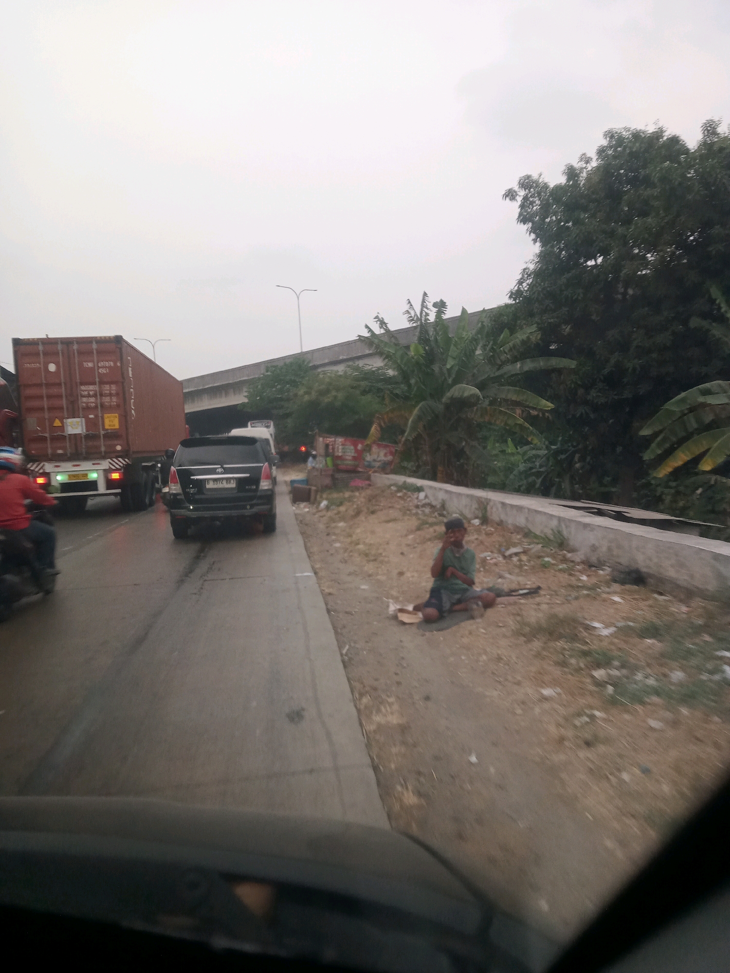 The image captures a scene on a busy road in Tangerang, Indonesia. A large container truck is prominently featured on the left, with a black SUV following closely behind. A motorcycle rider in a red shirt is also visible in the foreground. On the right side of the road, a young boy is sitting on the ground, appearing to be begging. He is wearing a green t-shirt and dark shorts. The environment consists of a paved road, a dirt shoulder, and lush green trees in the background, suggesting an urban or semi-urban setting. An elevated road or bridge structure can be seen in the distance. The sky is overcast, creating a diffused light.