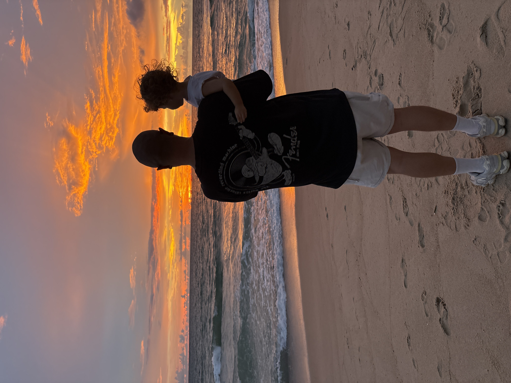 The media file depicts a father and son on a sandy beach at sunset. The father, with a shaved head, wears a black t-shirt and light-colored shorts. He is facing away from the camera, looking out at the ocean. The son, with curly hair, is being held by the father, resting his head on the father's shoulder and looking towards the sky. The son wears a white t-shirt.
The scene is a beach with waves gently rolling onto the shore. The sky is dominated by a vibrant sunset, with hues of orange, yellow, and pink illuminating the clouds. The sand has footprints scattered across it. The overall mood is serene and peaceful, suggesting a moment of family connection and appreciation for nature. The time of day is clearly sunset, characterized by the warm, golden light. The location cues, such as the sand and the ocean, confirm it is a beach setting.
Visible text on the father's t-shirt includes the word "Fender" and "since 1958", with some additional imagery that appears to be related to music or a record player.