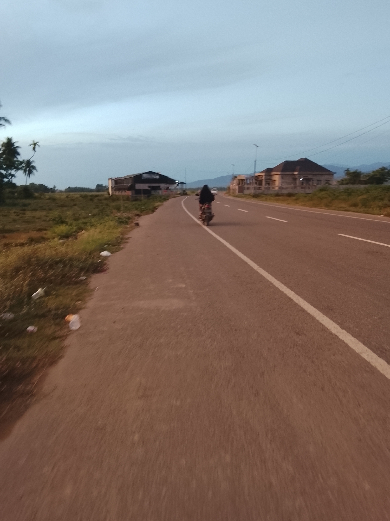 An image taken from a moving vehicle shows a two-lane asphalt road in Dayah Kruet, Indonesia, during twilight. The road is lined with a grassy shoulder and scattered debris. In the distance, several buildings are visible, including what appears to be a large, dark-colored structure on the left and a more modern, light-colored building with a dark roof on the right. A single person on a motorcycle is traveling away from the camera down the right lane of the road. The sky is overcast with a soft, diffused light. The general environment suggests a semi-rural or suburban setting along a main thoroughfare.
