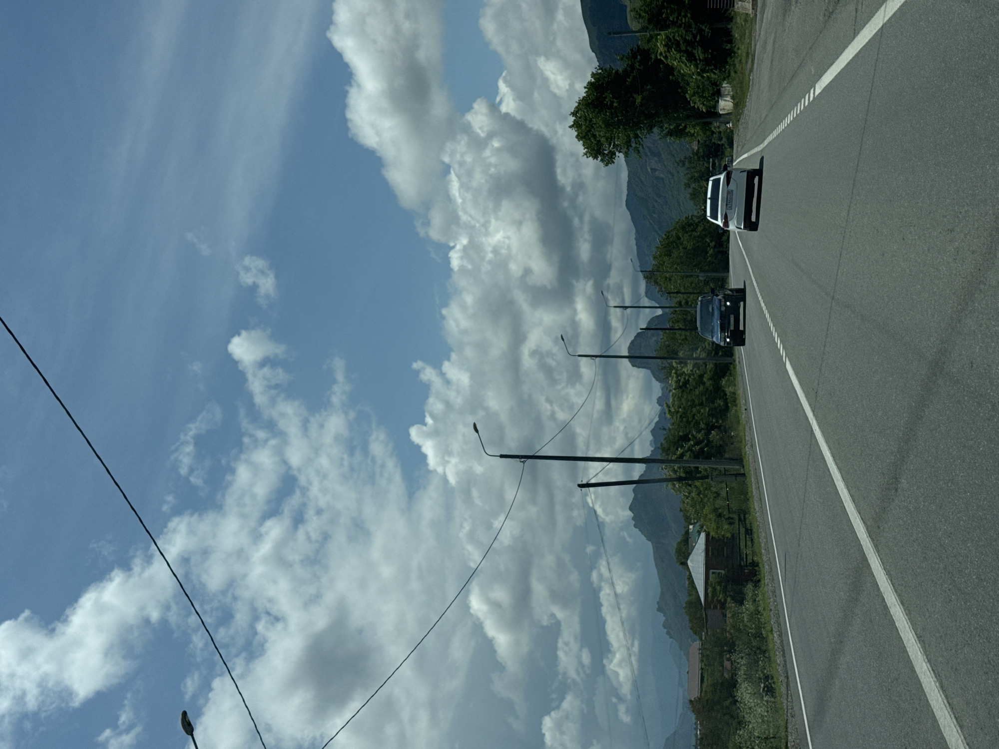 The image depicts a two-lane asphalt road with white lane markings. Two cars are traveling on the road: a dark SUV in the left lane and a white sedan in the right lane. Both vehicles are moving away from the viewer. The road is bordered by sparse vegetation, trees, and some buildings on the left. In the background, a range of mountains under a partly cloudy sky is visible. The sky is a light blue with scattered white cumulus clouds. The scene is illuminated by daylight. Based on the context of Красногорская, Russia, this appears to be a rural or semi-rural road leading towards mountainous terrain.