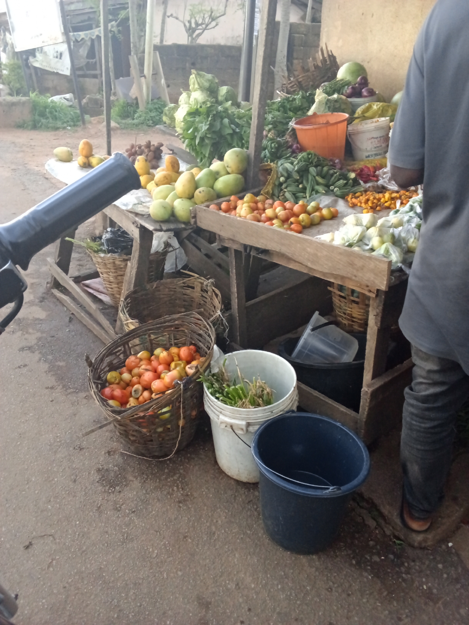 The image shows an outdoor market stall in Gura Top, Nigeria. The stall is laden with a variety of fresh produce, including tomatoes, mangoes, leafy greens, and okra. There are several woven baskets and plastic buckets used to display and store the goods. A person, partially visible from the chest down, stands at the right side of the frame, appearing to be a vendor or customer. The ground is paved with asphalt. In the background, there are buildings and vegetation. The overall impression is of a typical local market scene.