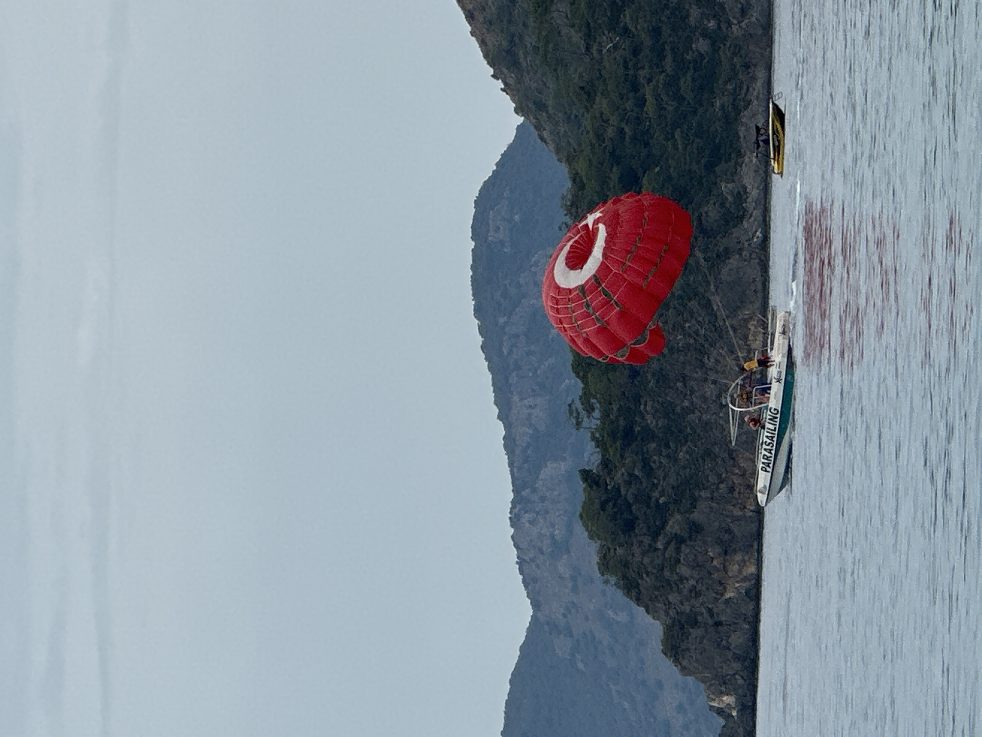 A landscape image shows a parasailing adventure against a scenic background. A red parachute adorned with a white crescent moon and star, reminiscent of the Turkish flag, hovers above a boat on the water, suggesting parasailing activity. The boat, labeled "PARASAILING", is likely in motion, towing the parachute. In the background, an imposing mountain range covered in lush greenery dominates the scene. The sky is overcast, casting a soft, diffused light over the landscape. This scene is likely located in Fethiye, Turkey, given the cultural symbolism on the parachute.