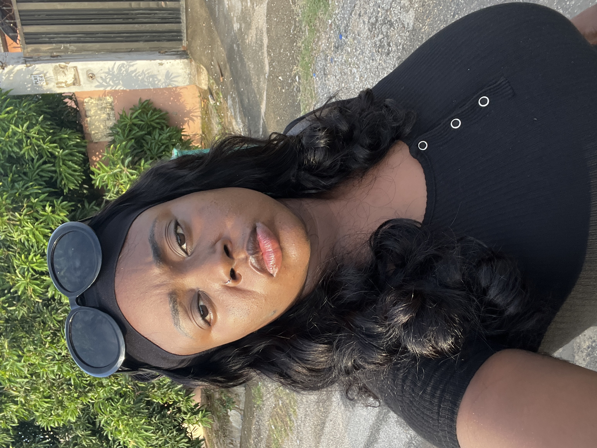 A young, dark-skinned woman is shown in a close-up selfie shot outdoors. She is wearing a black headband with dark, round sunglasses on top and a black, ribbed top with three visible white buttons. Her long, black hair is styled in loose curls. The background includes green foliage on the left and a building facade on the upper left. The ground is a mix of concrete and stone. While the specific location in Abuja, Nigeria is not discernable from the image, the presence of greenery and building styles are indicative of a residential or urban environment. There are no visible interactions or actions, as the subject is solely focused on the camera.