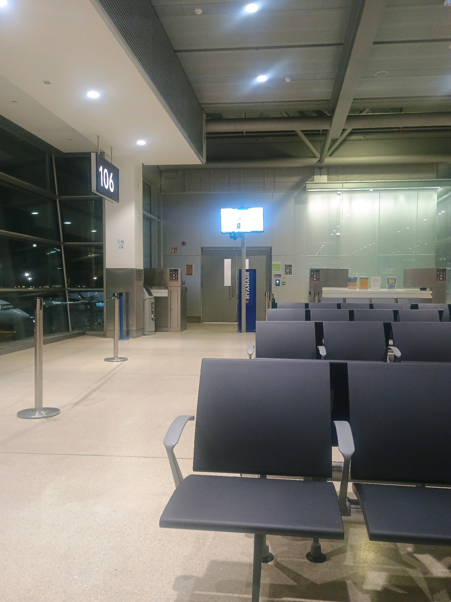 This image shows an empty airport departure lounge. In the foreground, rows of dark gray chairs are arranged neatly. To the left, a large window overlooks an airport tarmac at night, with an airplane visible. A digital sign displays "106" in white lettering against a black background, hanging from the ceiling. Further back, there are more chairs, a digital screen displaying flight information, and several service counters with brochures. The floor is light-colored and speckled. A blue stanchion with a retractable belt is positioned near the window. The overall environment is clean and functional, typical of an airport waiting area.