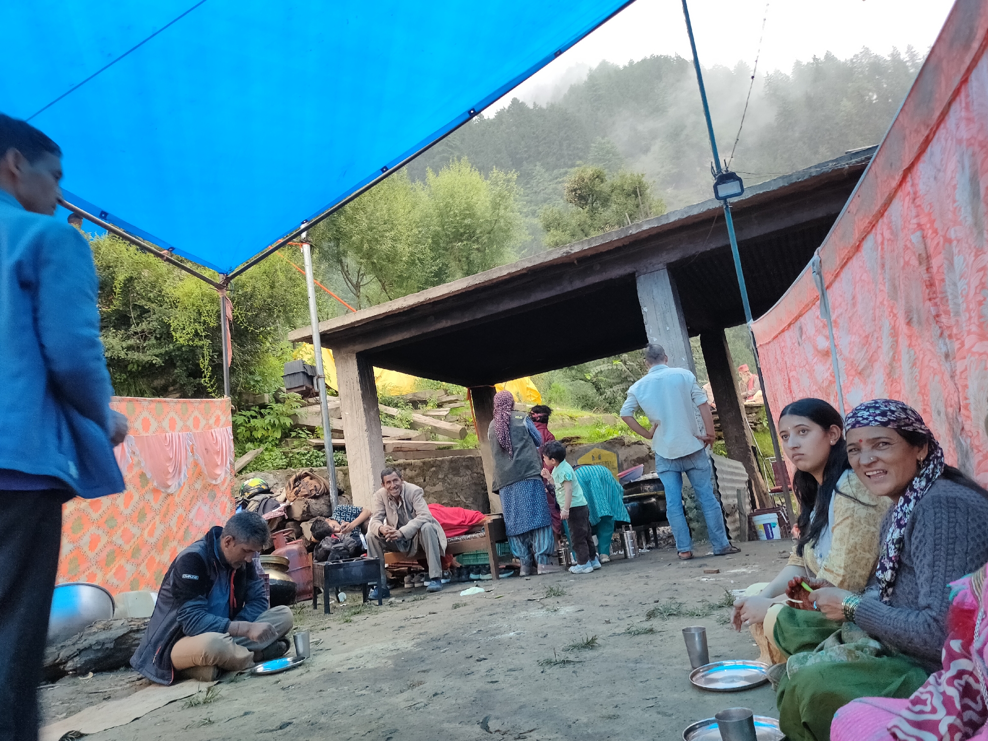 The image depicts an outdoor scene in Damadi, India, likely a rural or mountainous area, under a large blue tarpaulin shelter. Several people are gathered, some sitting on the ground and others standing. In the foreground, a man in a dark jacket and tan pants is seated on the dirt, eating from a plate. To his right, two women are seated; one in a yellow outfit looks towards the left, while the other in a grey sweater and patterned headscarf is smiling and holding a green object. Behind them, a group of people, including children, are interacting near a simple structure with a concrete roof. The environment features a backdrop of lush green trees and misty mountains, suggesting a hillside location. Various objects are scattered around, such as cooking pots, a grill, and what appear to be camping supplies, indicating a gathering or a temporary settlement. The lighting suggests it is daytime.