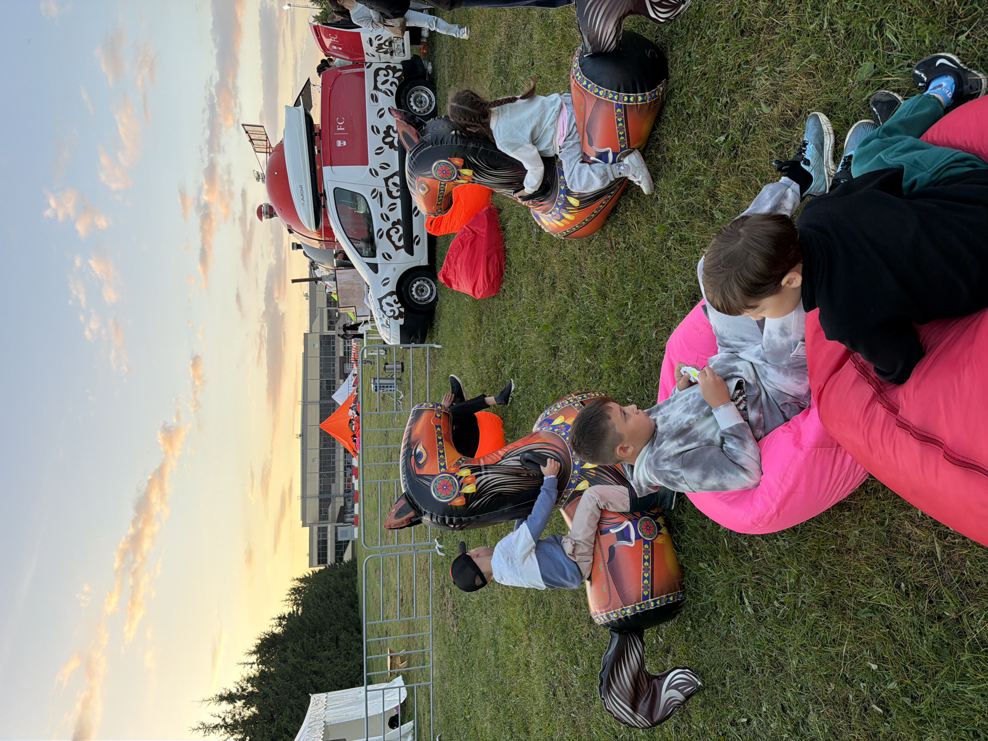 In a grassy outdoor area, likely at dusk, children are engaged with inflatable horse toys and beanbag chairs. One young boy, wearing a grey tie-dye hoodie and grey sweatpants, sits on a bright pink beanbag chair, looking at a small device. Another boy in a black long-sleeved shirt and green pants sits on a red beanbag chair. A third child, a girl with braided hair, is seated on an orange and black inflatable horse toy. To the left, a boy wearing a black baseball cap, a blue and white long-sleeved shirt, and tan pants rides a similar inflatable horse.
In the background, a white van with red accents and "FC" branding is parked, and beyond that, a large building with a flat roof is visible. Several orange and white tents are also present in the distance, along with a row of coniferous trees. The sky is partly cloudy with hues of orange and pink, suggesting sunset. A metal fence runs across the middle ground.