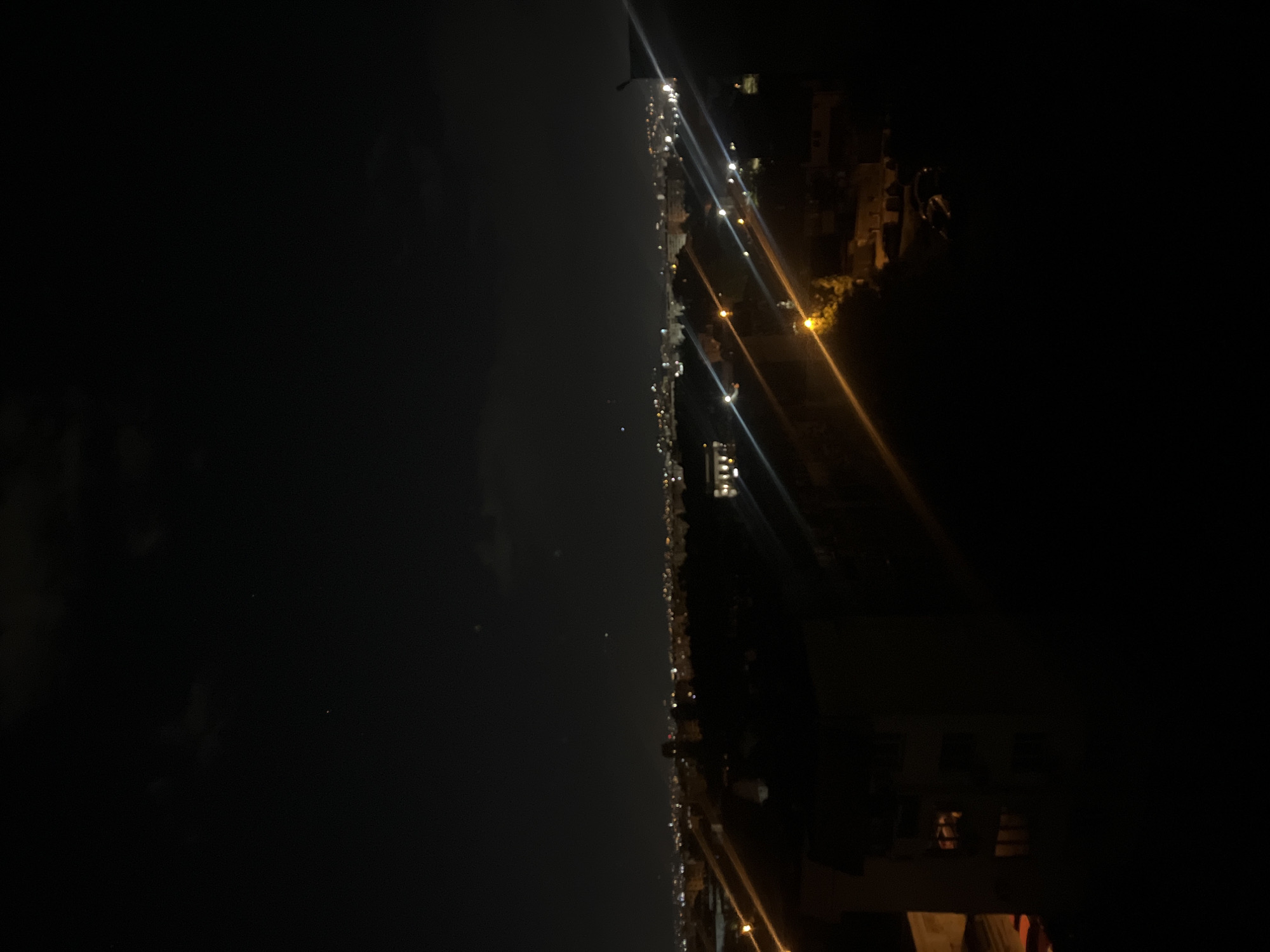 The image depicts a nighttime view of a city, likely from an elevated position. A dark sky dominates the left side of the frame. The city's illuminated coastline stretches towards the right, revealing streetlights and buildings. There are no people visible, and the subjects are primarily the urban landscape and night sky. Based on the location, it is not possible to confirm that it is the city of Sofia.