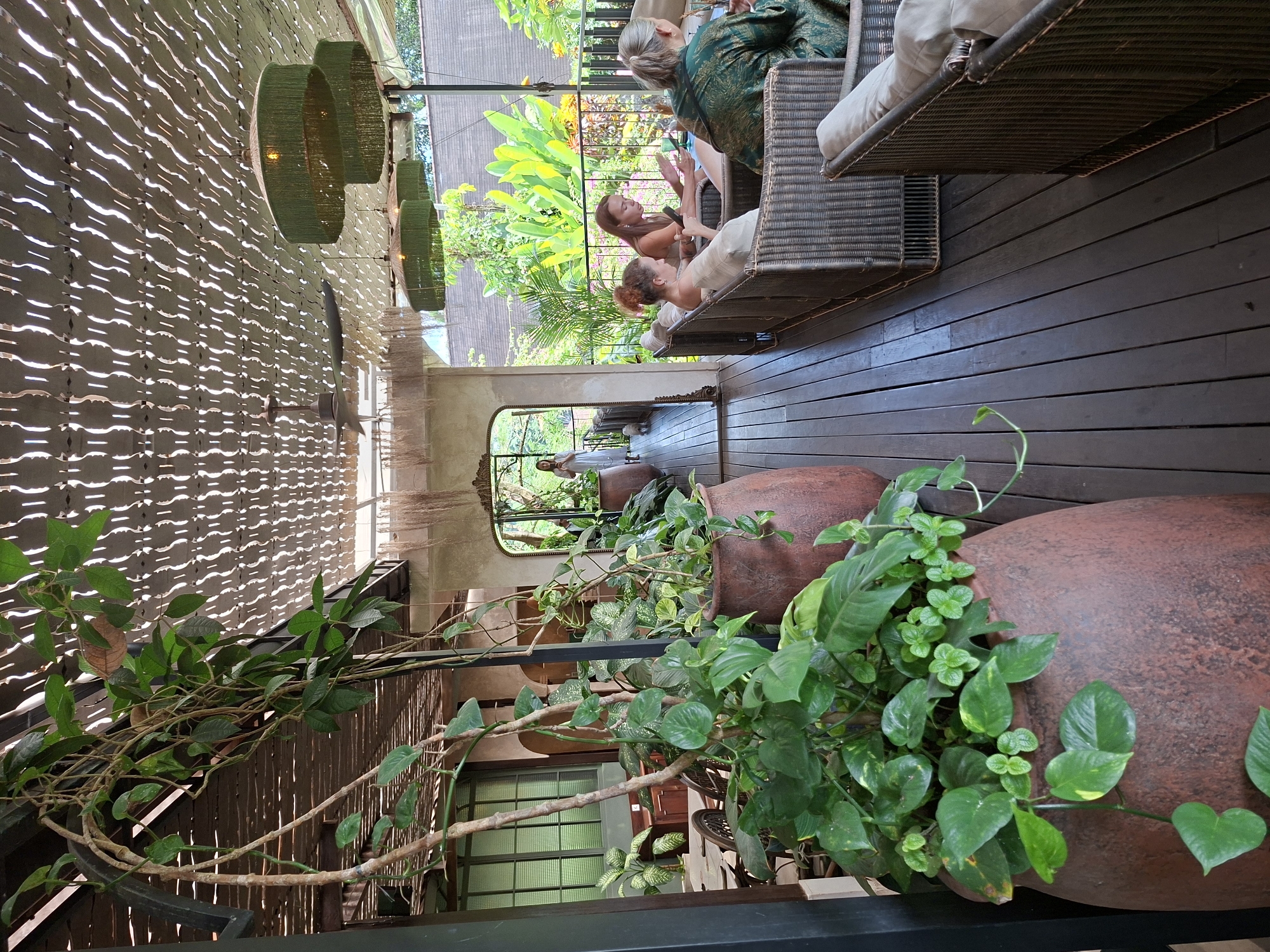 The image captures an indoor-outdoor seating area in the Ubud District of Indonesia. Two individuals are seated on wicker chairs, appearing to converse. Adjacent to them, another person is also seated.
In the foreground, large terra cotta pots filled with green plants take prominence. These pots sit on a platform behind a black metal railing. The flooring is dark wood, running horizontally across the frame.
Overhead, a wooden awning with patterned cutouts casts light and shadow. Green hanging lamps hang down. In the background, an arched doorway reveals a figure in white walking through. The backdrop contains lush greenery, suggesting an open or outdoor space beyond the seating area.