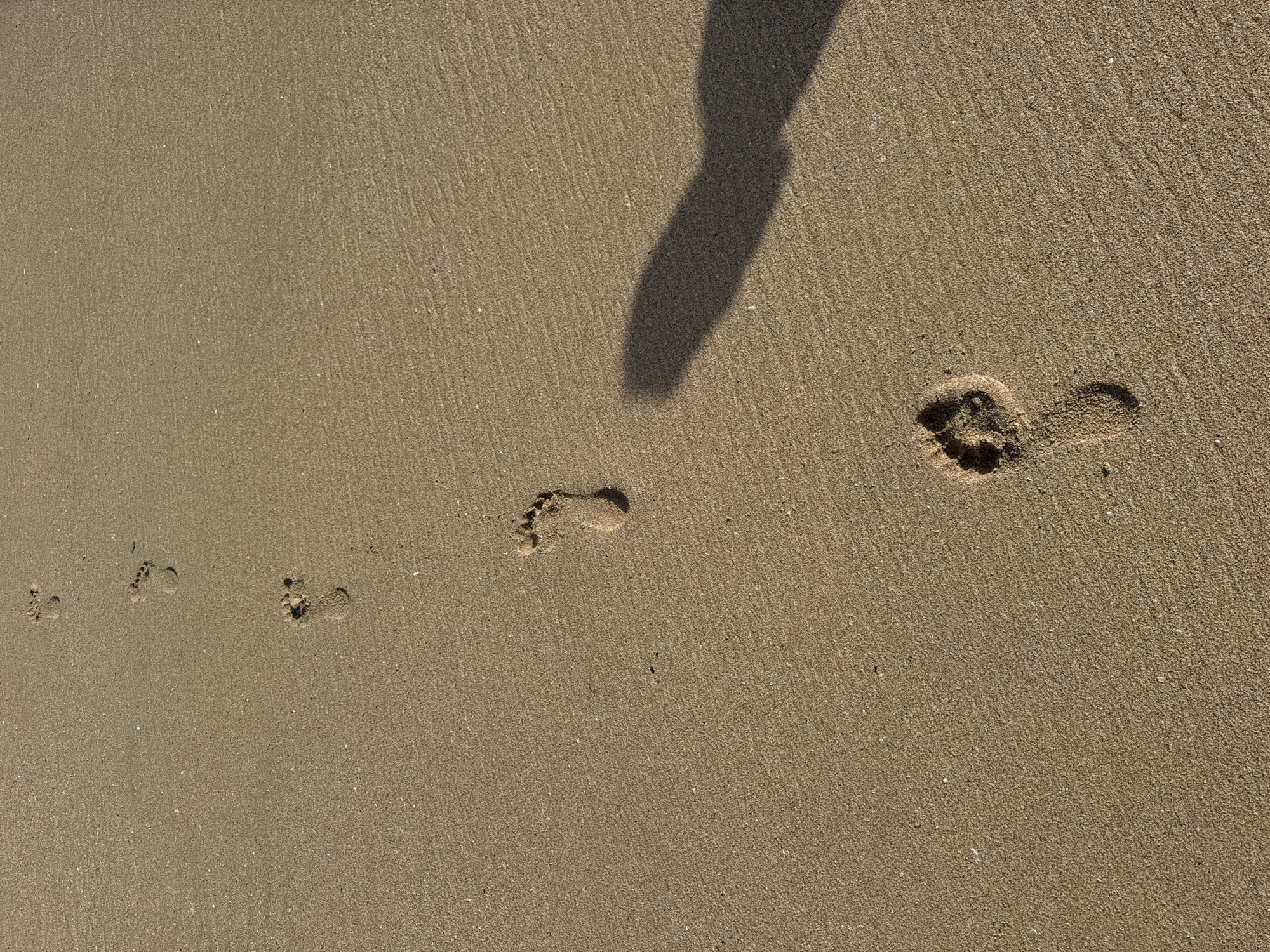 The scene is a sandy beach in Kuta Selatan, Indonesia. The sand is fine and light brown in color. There are a series of barefoot footprints leading from the lower left of the image towards the upper right. The prints vary in clarity, with some being more distinct than others. In the upper right portion of the image, a dark shadow is visible, likely cast by a person standing nearby.