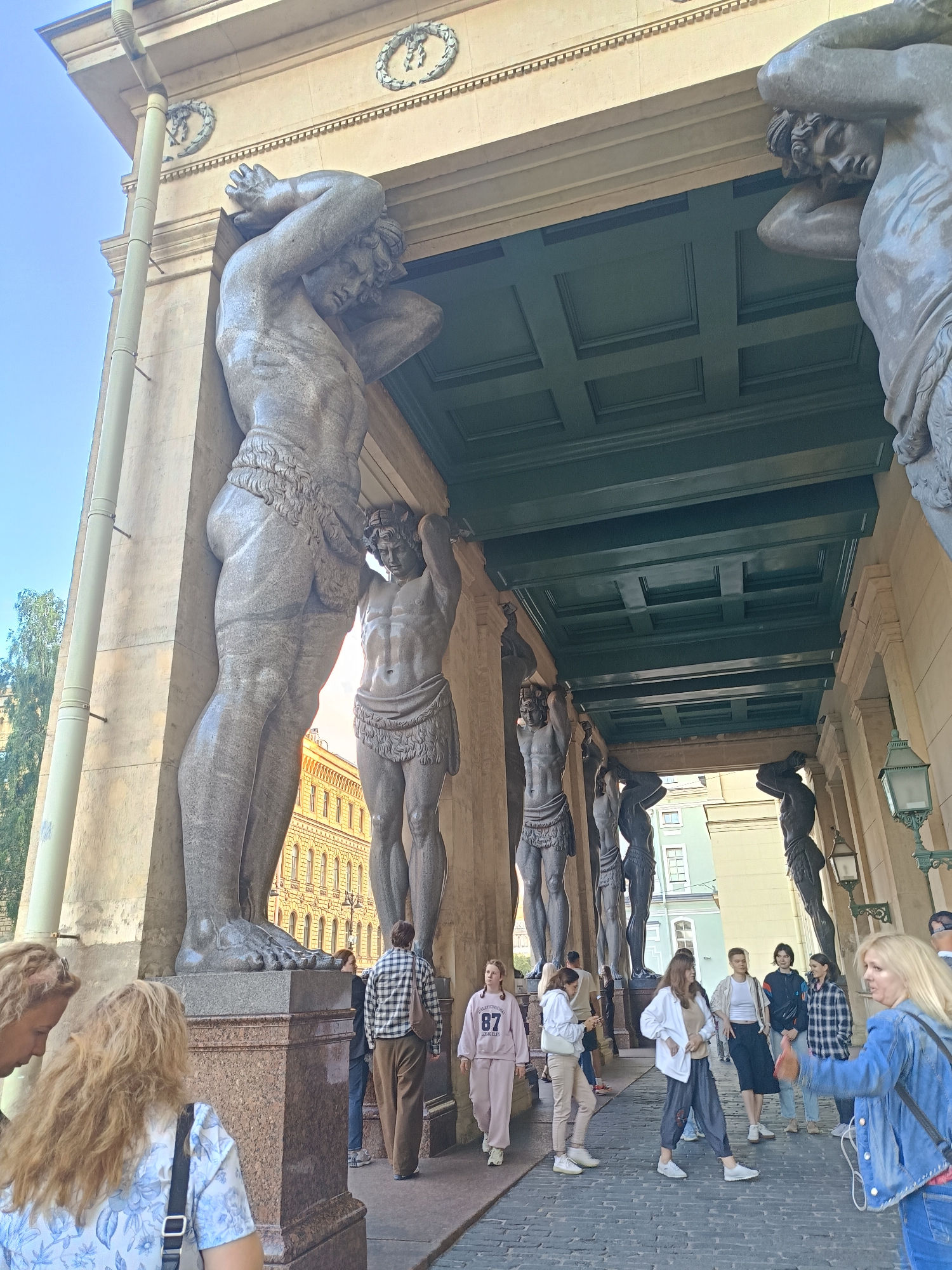 In the city of Unknown, Unknown, a group of people are gathered under a covered walkway supported by large, stone statues of men holding up the structure. The statues are muscular and depicted in a classical style, with some wearing loincloths. The walkway has a green, paneled ceiling. A building with arched windows is visible in the background. The people in the foreground and midground are diverse in age and gender, dressed in casual clothing. Some are walking, while others appear to be standing and observing the statues. One woman in the foreground on the right is wearing a denim jacket and jeans and appears to be speaking to someone off-camera. The ground is paved with cobblestones. The lighting suggests it is daytime, with sunlight casting shadows.