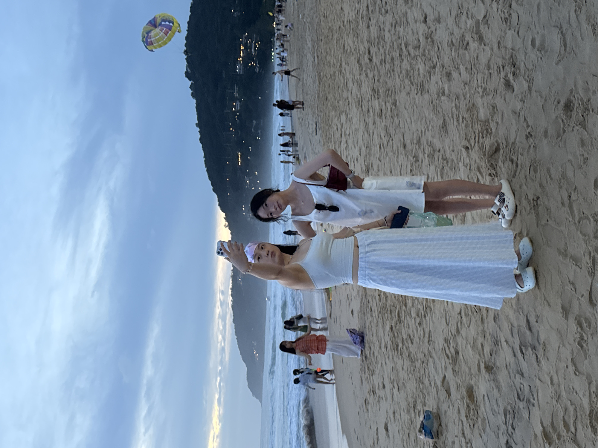 Two young Asian women are taking a selfie on a sandy beach. The woman on the left is wearing a white tank top and a long white striped skirt. She has a light pink headband on and is holding up her phone to take a picture. The woman on the right is wearing a white sleeveless dress and has a dark red purse hanging from her shoulder. They are standing on the beach with the ocean and waves in the background. In the distance, there is a hill covered in trees and lights, and a yellow and purple parachute is visible in the sky. Other people are scattered along the beach and in the water. The sky is cloudy and the lighting suggests it is either early morning or late evening. The location is Karon Beach in Karon, Thailand.