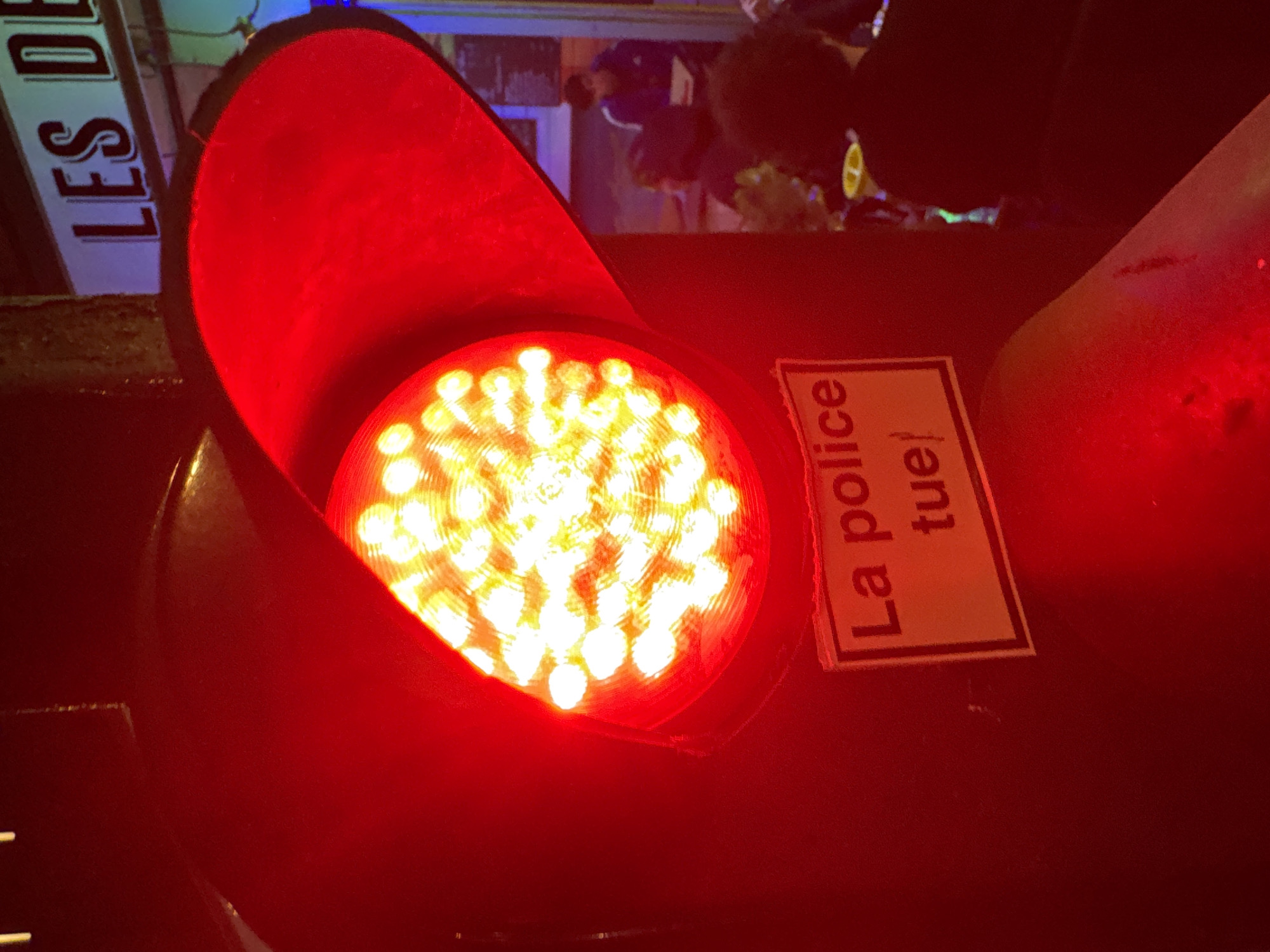 The image captures a close-up, angled view of a red LED traffic light illuminating an urban night scene in Paris, France. The traffic light, prominent in the foreground, glows intensely red, casting a warm hue across much of the frame. The light source consists of numerous individual LED bulbs arranged in a circular pattern, visible through the lens.
Attached to the traffic light pole, to the right of the red signal, is a white rectangular sticker with black text. The text clearly reads: "La police tue!" followed by an exclamation mark, which translates from French to "The police kill!".
In the blurred background, several elements are discernible. To the left, a partial sign with white lettering on a dark background is visible, showing the text "LES DE". Further back, indistinct figures of people can be seen, suggesting an active street environment. One figure on the right appears to have a yellow object or detail, possibly part of a bicycle wheel, near them. The overall setting implies a street or intersection in a city at night or dusk, emphasized by the artificial lighting and the dark, indistinct surroundings. The strong red light from the signal dominates the visual tone of the image.