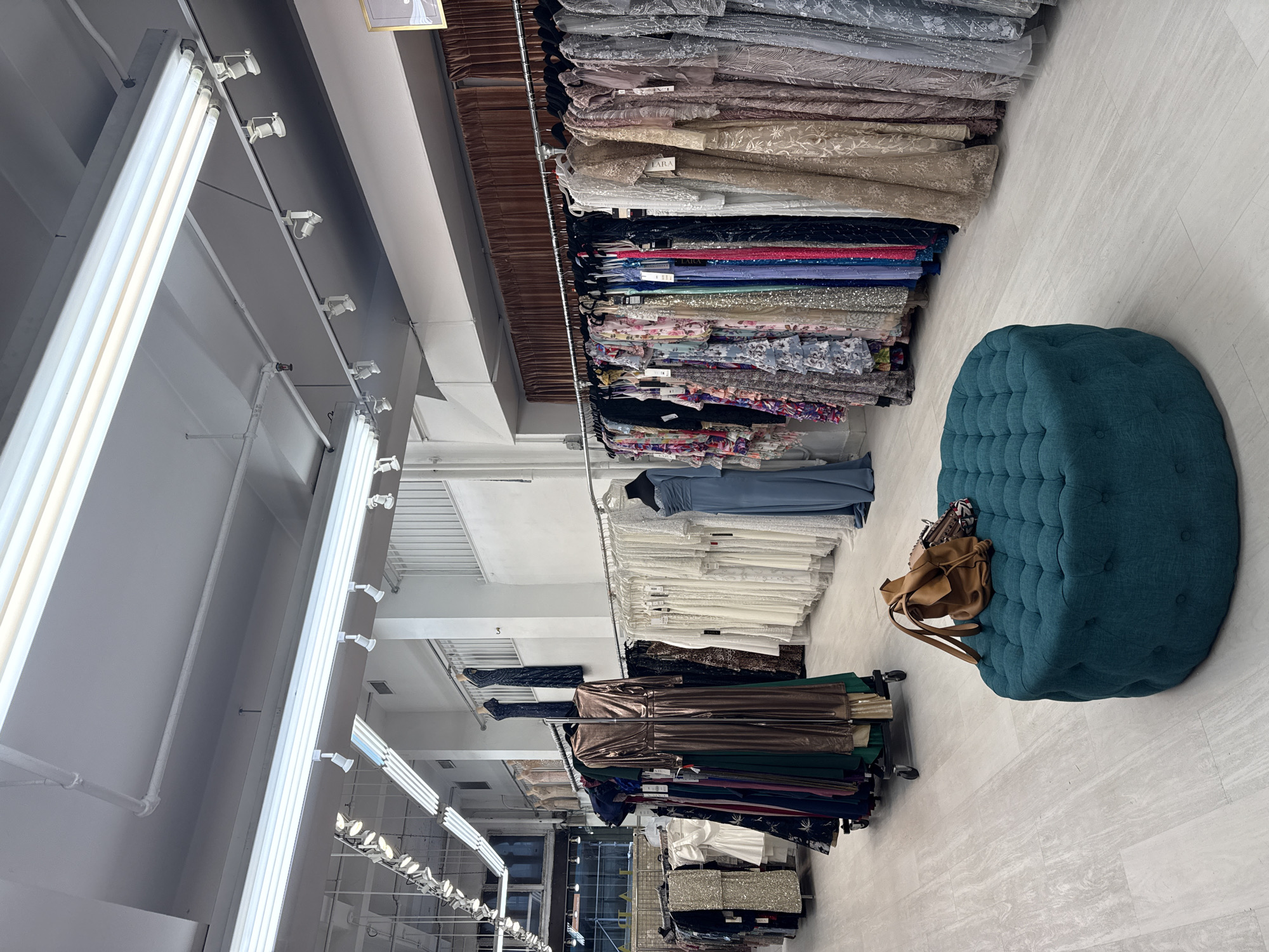 This is an interior shot of a dress boutique in New York City, United States of America. The store features racks of formal dresses, predominantly in various colors and styles, hanging from silver racks. A large, teal tufted ottoman sits in the foreground, with a brown leather backpack and a smaller purse resting on it. The floor is made of light-colored wood-look tiles. The lighting appears to be from fluorescent and track lighting fixtures on the ceiling. There are no people visible in the image.