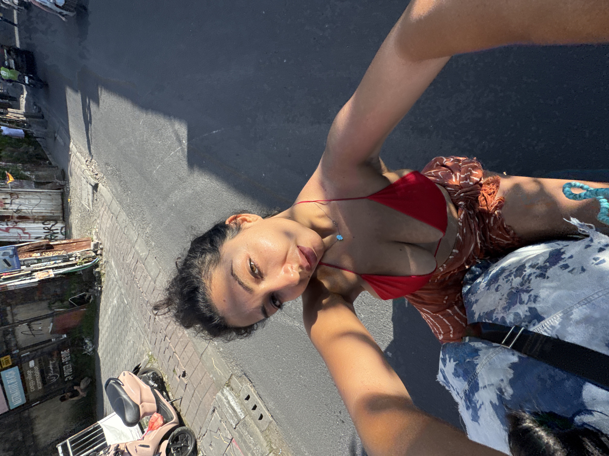 A selfie taken at street level shows a young woman in a red bikini top and patterned skirt standing on an asphalt road in North Kuta, Indonesia. In the lower left, a pink mobility scooter is parked near a building with signage, including "POWER-NERVE." A person is visible near the building entrance. Across the street, various objects are piled against a building. The road stretches away in the background. The woman's left arm and a portion of what appears to be a tie-dyed blue and white bag with a black strap are in the lower right foreground.