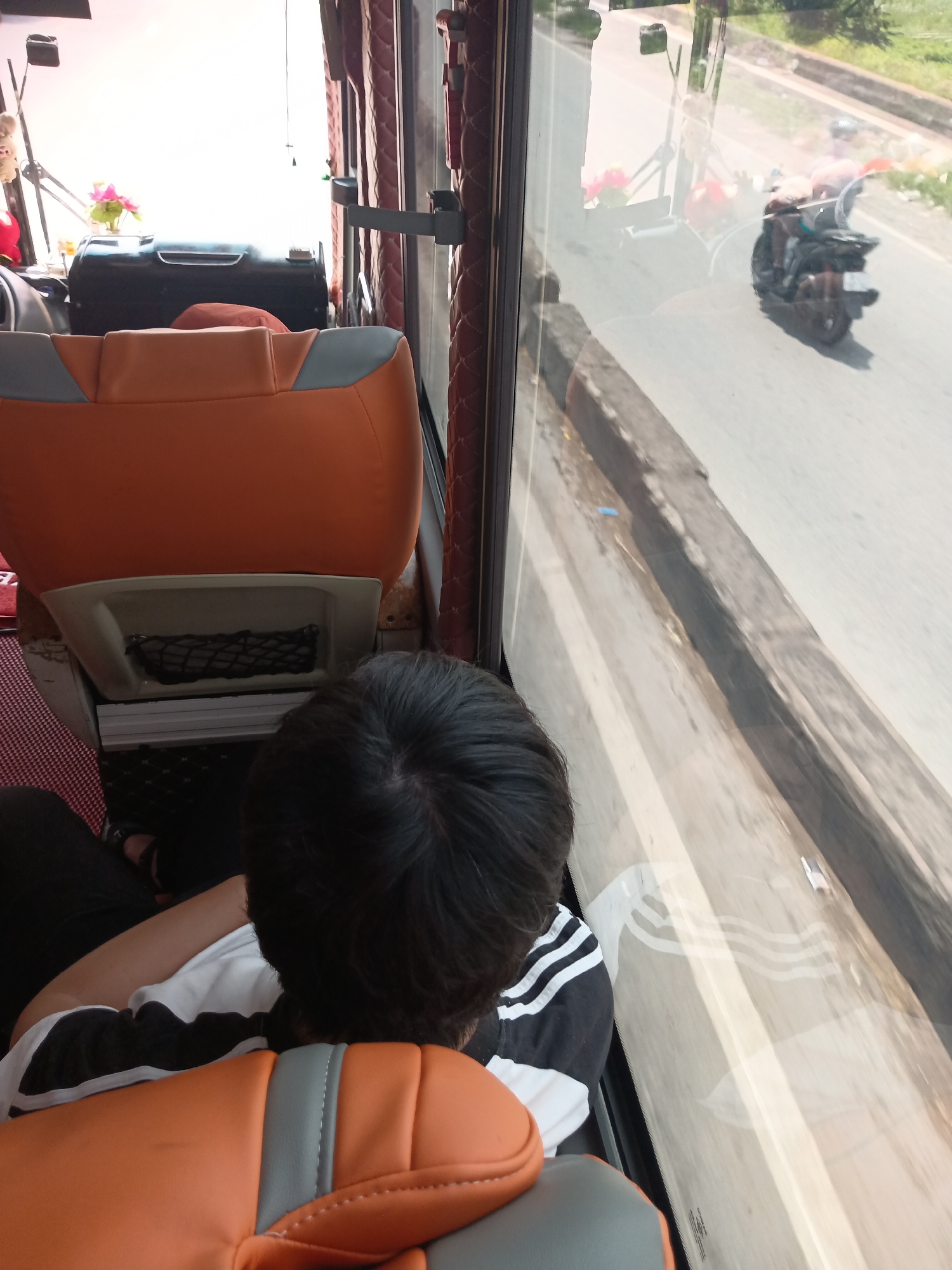 A person is seated inside a vehicle, looking out the window. The interior of the vehicle appears to be a bus or a large van, with orange and gray cushioned seats visible. A black storage compartment with a net is attached to the back of the seat in front. Outside the window, a road is visible with a motorcycle riding in the distance. The environment outside appears to be a sunny day in an urban or suburban setting.