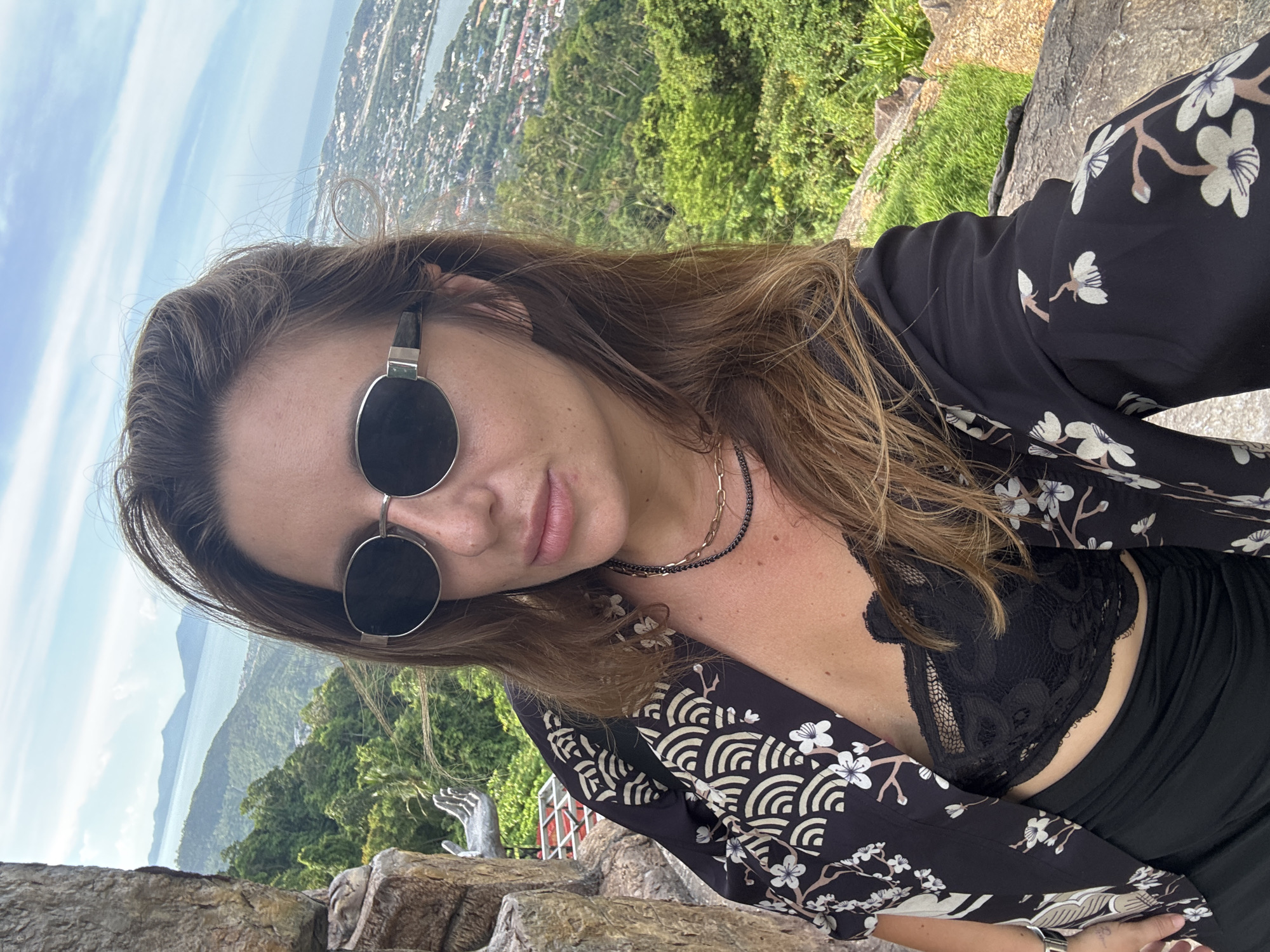 A woman is taking a selfie outdoors with a scenic view of a coastal city and mountains in the background. She is wearing sunglasses, a black lace bralette, a black open-front shirt with a floral and wave pattern, and a black skirt. She is standing next to a rock formation. The location appears to be a viewpoint overlooking a city and a body of water.