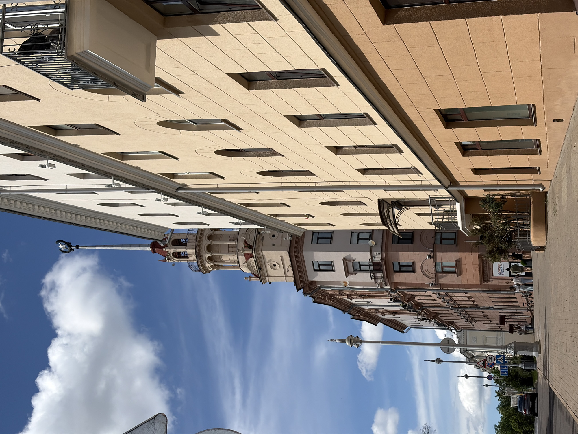 This image depicts a sunlit street scene in Minsk, Belarus, specifically featuring the "Building with a Spire" (also known as the House of Officers or House with the Star) at Kirova Street 11.
The prominent structure is a multi-story building on the left and center, constructed in a light beige and cream color, characterized by Stalinist Empire architectural style. It features a tall, ornate corner tower with a red-tiled conical roof supporting a slender white spire. The spire is topped with a white five-pointed star enclosed within a wreath. Below the spire, the tower includes a circular balustraded observation deck with classical columns. The building's facade displays rows of both rectangular and arched windows. An adjacent section of the building to the left is a muted pinkish-brown, also featuring multiple windows and small balconies.
To the immediate right, a light yellow-beige multi-story building rises, presenting a linear facade with distinct rows of arched and rectangular windows. A prominent balcony with a black wrought-iron railing protrudes from an upper floor of this building; a dark, indistinct object is visible on it. Below this, a section of a darker orange-brown building is present, featuring rectangular windows at street level.
The foreground is a clean, paved sidewalk made of light brown brick pavers arranged in a herringbone pattern. An asphalt road runs parallel to the sidewalk on the left. Several tall, grey, ornate lampposts with decorative finials line the sidewalk. Visible traffic signs include a "40 km/h" speed limit, a pedestrian crossing sign, and blue directional signs. Multiple individuals, predominantly adults, are observed walking on the sidewalk near the pinkish-brown building, with some congregating near a recessed entrance that displays a "SOCCER SHOP.BY" sign. A dark-colored sedan is partially visible on the road in the mid-distance.
The scene is illuminated by bright daylight, casting distinct shadows. The sky is a clear blue with scattered, fluffy white cumulus clouds. Security cameras are mounted on several building facades.