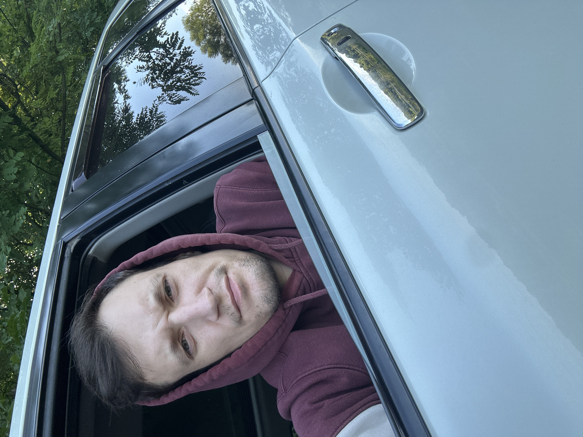A man is seen through the open window of a light blue car. He is wearing a burgundy hooded sweatshirt with the hood pulled up around his head. The man is looking directly at the camera with a neutral expression. The car's interior is dark, and the exterior is a light, metallic blue. The car door handle, a chrome rectangle, is visible on the right side of the image. Reflections of trees and the sky are visible in the car window. The background suggests an outdoor setting with green foliage. The image appears to be a selfie taken from inside the car. There are no visible markers or clues that indicate the specific location within Makiv, Ukraine.