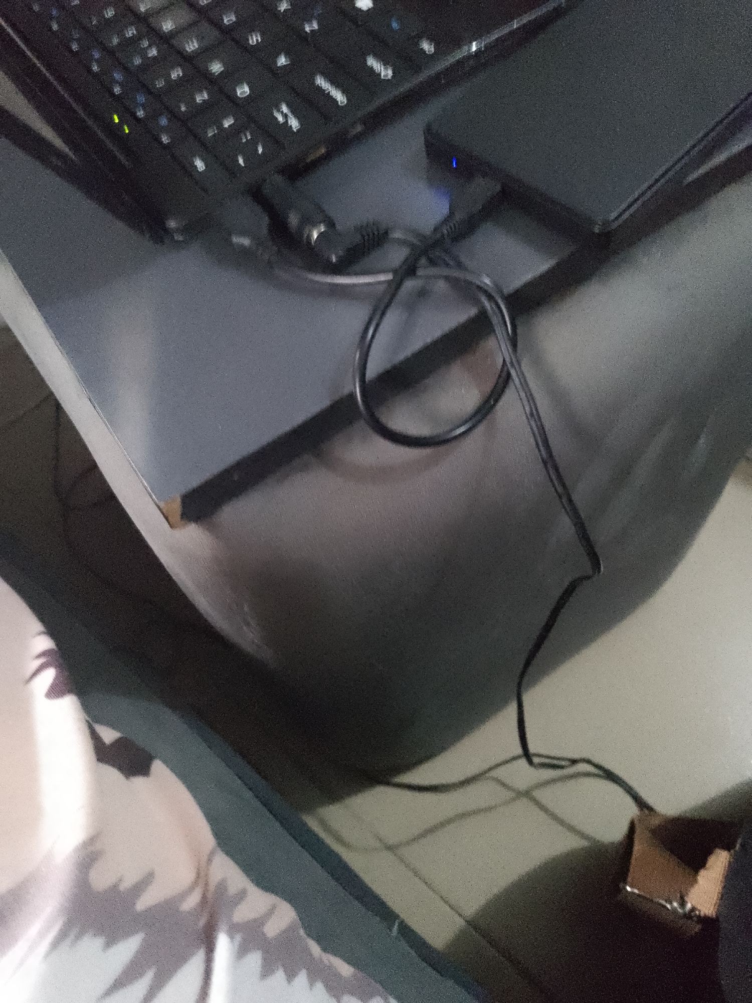 The image shows a close-up, blurry view of a laptop keyboard and a black external hard drive with a blue indicator light. A black cable snakes from the laptop to the hard drive and then continues to a small, damaged cardboard box on the floor. A portion of patterned fabric, possibly clothing, is visible in the lower left corner. The scene appears to be indoors, on a grey surface that could be a desk or table. The lighting is dim. There are no people visible in the image. The location in Jauro Yinu, Nigeria, cannot be determined from the visual information provided.