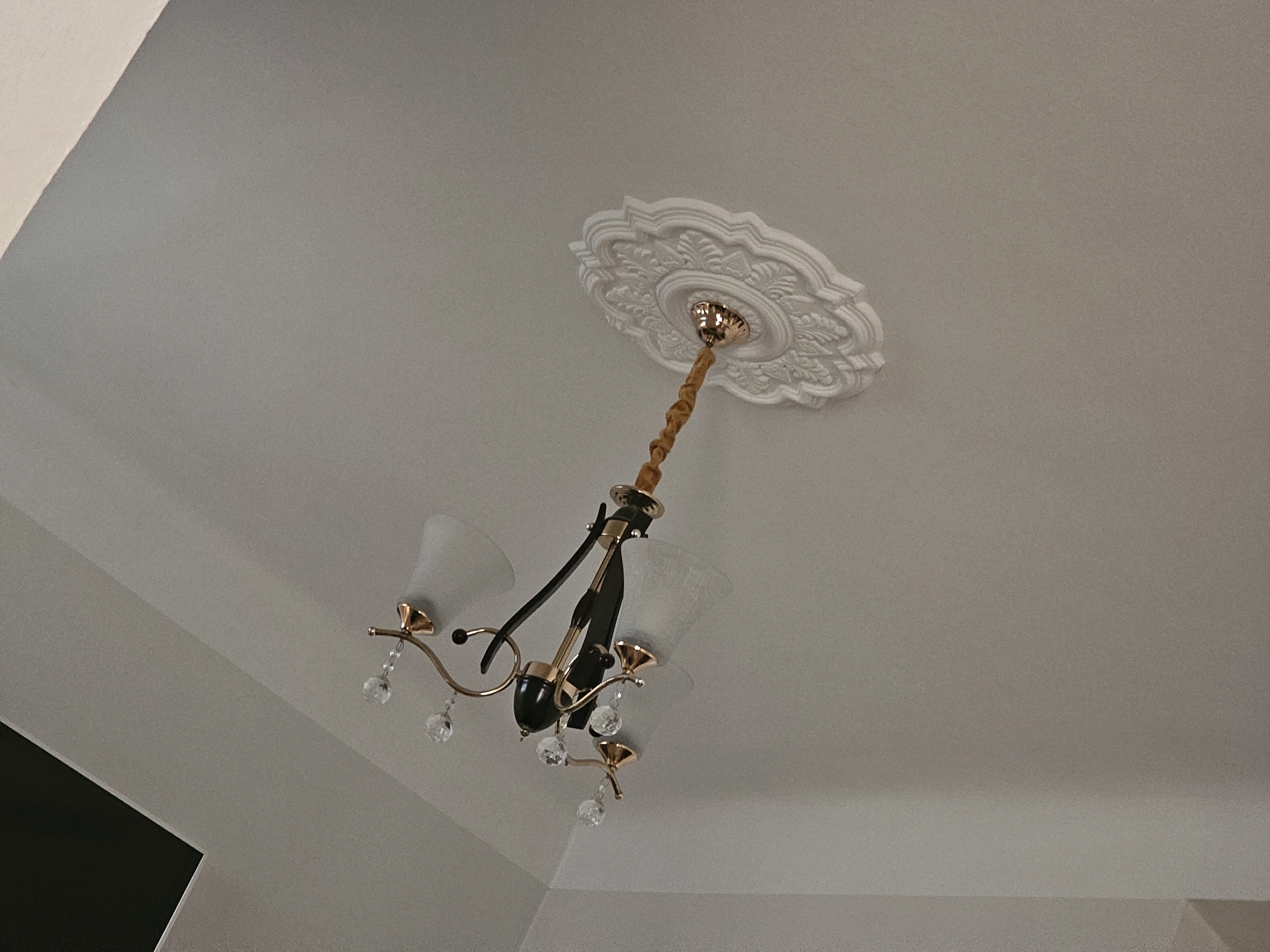 The image shows a chandelier hanging from a white ceiling with a decorative medallion. The chandelier has a gold-colored frame, frosted glass shades, and clear crystal accents. The ceiling appears to be in a residential setting in the city of Batna, Algeria. There are no people or visible actions in the image. The environment is indoors, with a plain white ceiling.