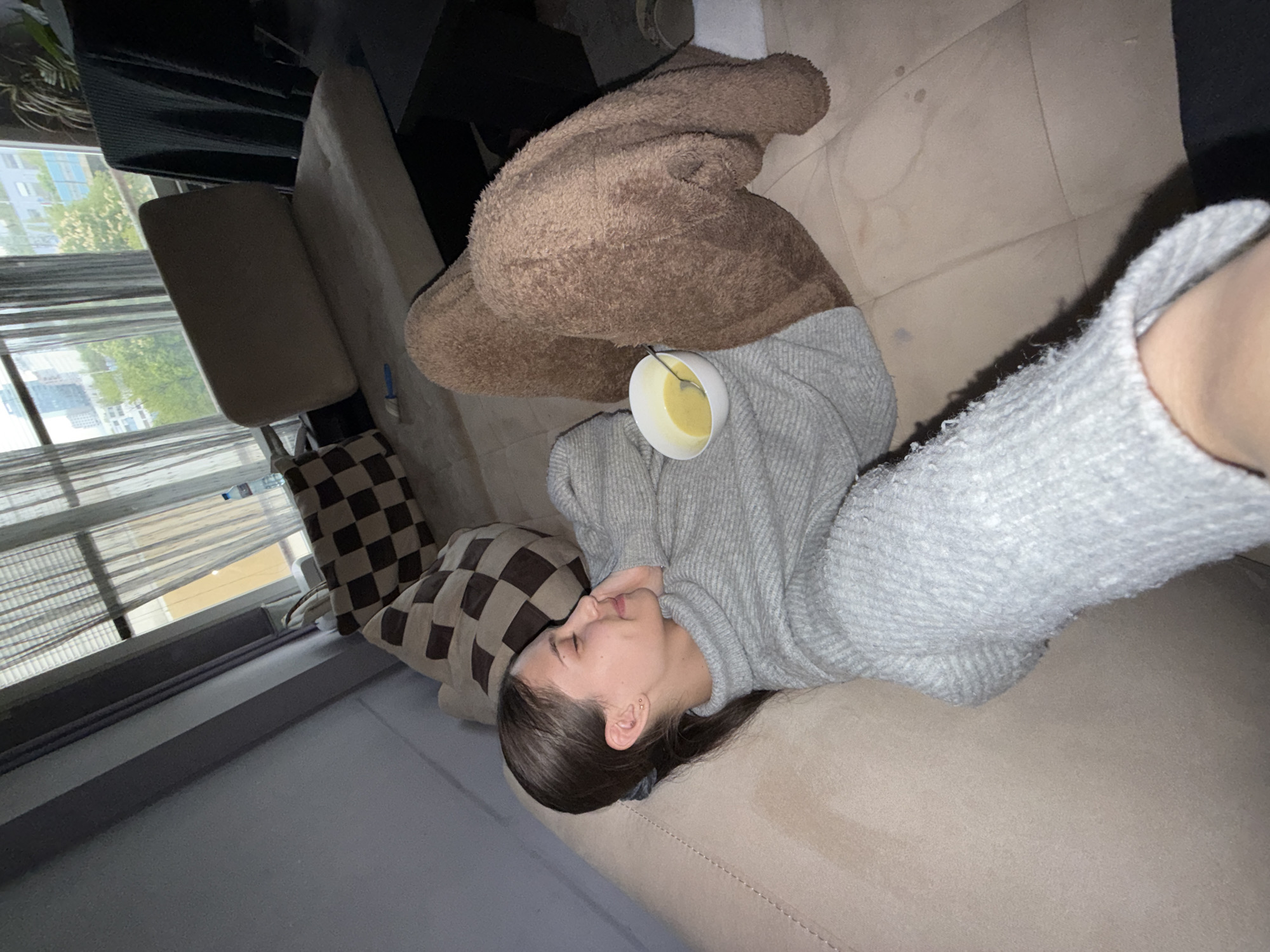 A young woman is depicted resting or sleeping on a light-colored fabric couch in an indoor setting in Sofia, Bulgaria. She is lying on her back, with her head turned to the left and her eyes closed, conveying a sense of peace and relaxation. Her dark hair is casually styled, and she wears a light grey, possibly knitted, long-sleeved top. Her lower body is covered by a fluffy, brown, plush blanket or loungewear.
A small white bowl containing a light yellow liquid, possibly soup or a beverage, along with a spoon, is carefully balanced on her stomach/chest area. A brown and off-white checkered pillow supports her head. To the left, a large window, partially covered by sheer vertical blinds or curtains, provides a view of an urban landscape with green trees and city buildings visible in the distance. The lighting is soft and ambient, suggesting daytime, possibly late afternoon. No visible text is present in the image. The scene conveys a moment of quiet rest in a comfortable home environment.