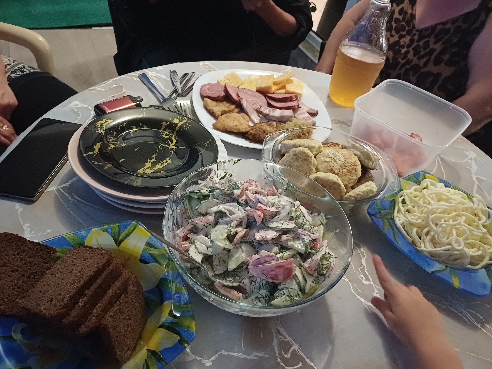 A top-down, close-up shot shows a table laden with food, possibly in Sochi, Russia. A person's hand reaches towards a plate of spaghetti on the right. A bottle of beer sits near a woman with a leopard-print top on the far right, her arm resting on the table. Another person, partially visible in the background, wears a black shirt. The table is covered with a grey and white patterned tablecloth. Food items include a salad in a glass bowl, a plate of sliced bread and a black and gold patterned plate with stacked dishes on the left. In the center, a white plate holds a selection of sliced meats, cheeses, and what appear to be fried pastries. Another glass bowl contains round, flat food items, possibly patties or flatbreads. A small red electronic device with the brand name "JUUL" is visible on the left side of the table, near a smartphone. The lighting is ambient, suggesting an indoor setting.