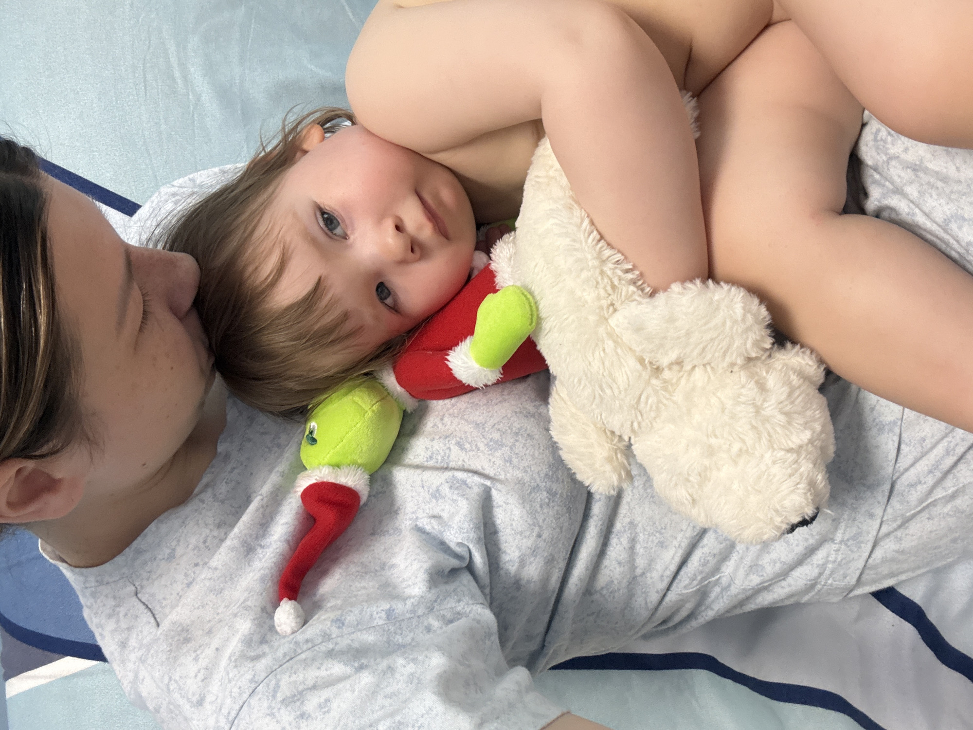 A woman and a young child are lying on a light blue bed with dark blue stripes. The woman, on the left, is wearing a light blue and white patterned shirt and has her eyes closed, appearing to kiss the child's forehead. The child is positioned on the woman's torso, partially undressed, and holding a white plush dog toy and a green Grinch toy. The child is looking towards the camera. The image is taken from an overhead perspective. The location is unknown.