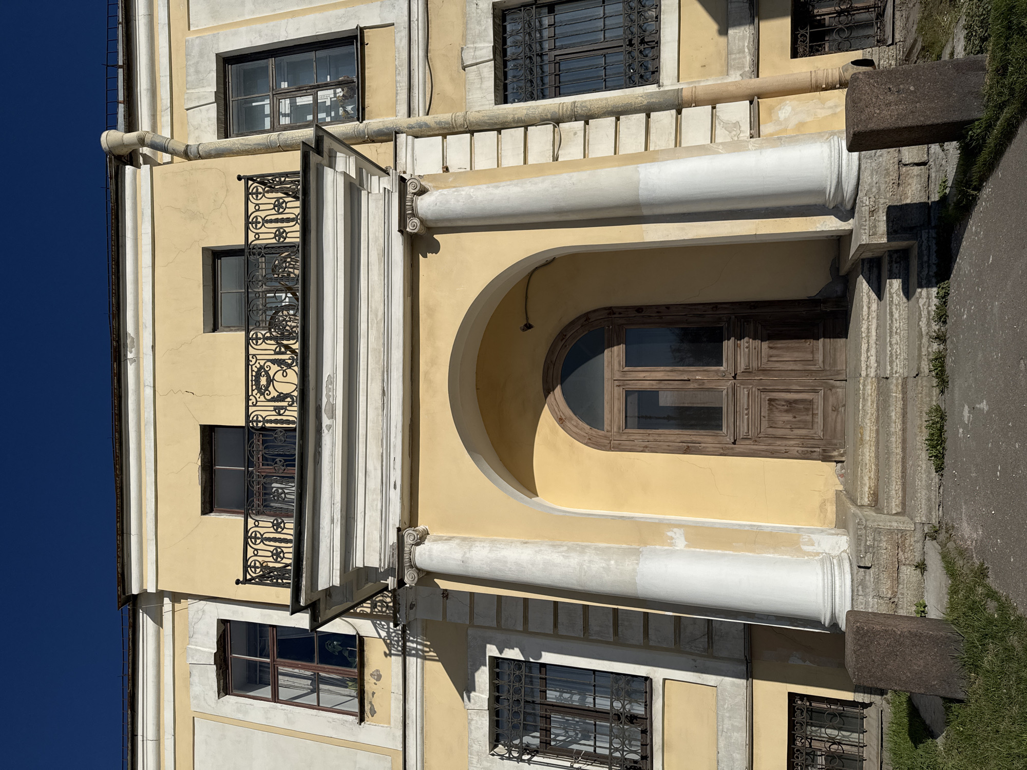 The image shows the facade of a building in Saint Petersburg, Russia. The building is yellow with white architectural details, including columns and decorative moldings. There is a dark wooden door with an arched top, flanked by white columns. Above the door is a small balcony with an ornate wrought-iron railing. The building has multiple windows with dark frames and metal bars. A gutter runs along the top of the facade. The sky is visible on the left side of the image and is a deep blue. The photograph is taken from a ground-level perspective and appears to be oriented vertically, with the building tilted. The scene is lit by natural daylight.
