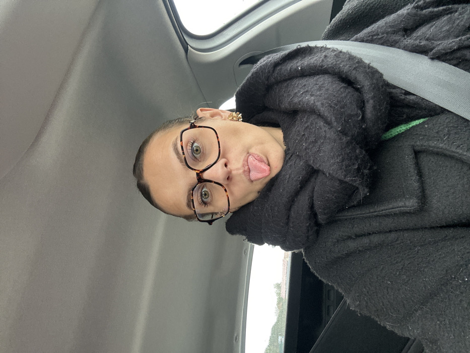 A female subject, an adult, is depicted in a close-up, front-facing photographic portrait taken inside a vehicle in Saint-Cyprien, France. She has her hair pulled back and wears large, tortoise-shell patterned eyeglasses. Her eyes are open, and she extends her tongue outwards in a prominent, expressive gesture. She is dressed in a dark, textured coat and a voluminous, heavily textured black scarf wrapped around her neck. A gray seatbelt is fastened across her chest. A gold-toned, ornate earring is visible on her right ear. The interior of the vehicle, including the light gray headliner, is visible, along with an overcast sky through the side and rear windows. Blurred, distant structures, consistent with residential buildings, are faintly visible through the rear window.