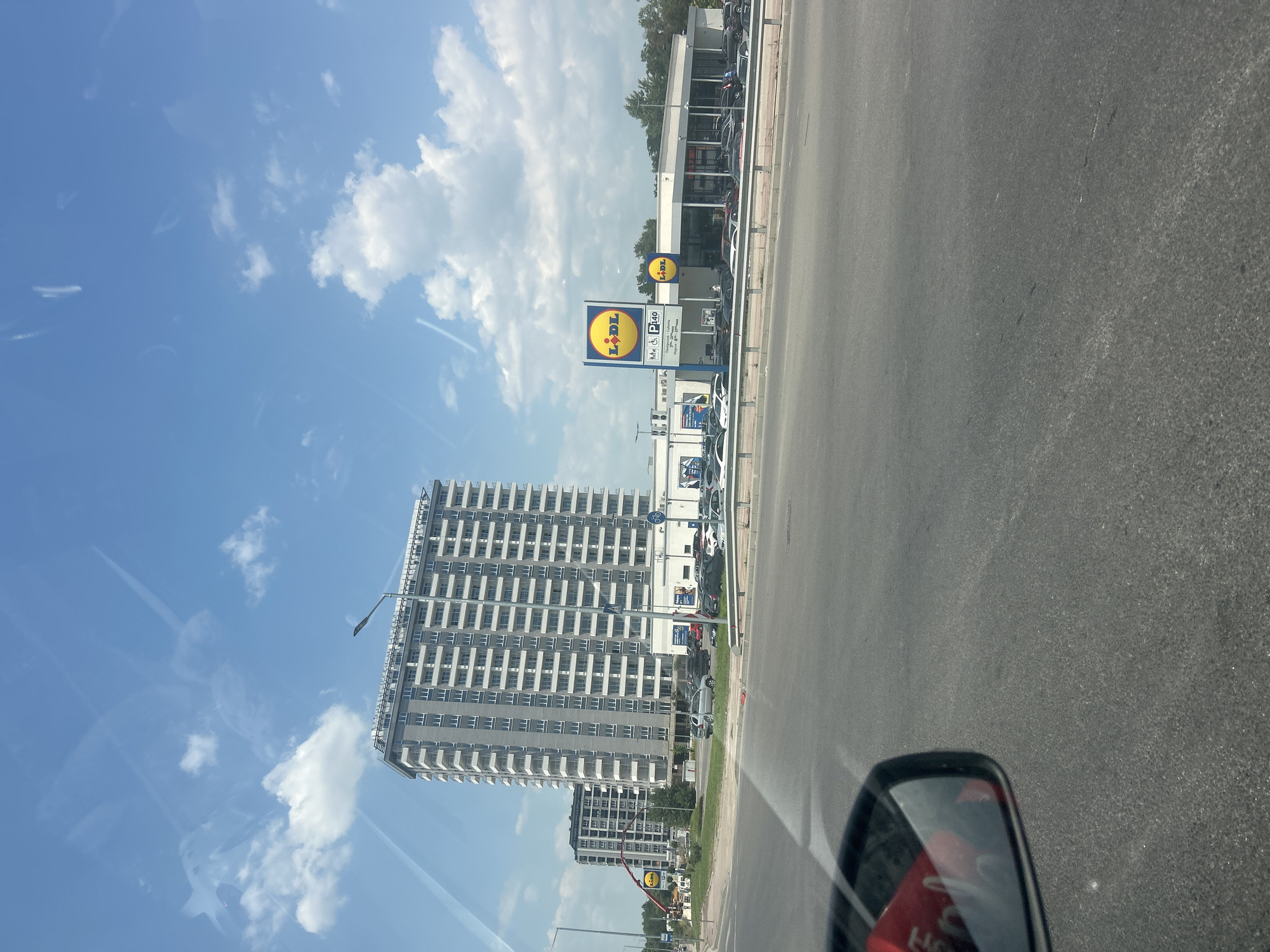 The image captures a street scene in Plovdiv, Bulgaria, on a bright, sunny day with scattered white clouds in a blue sky. The perspective is from a vehicle, looking slightly upwards and to the right. A prominent tall, modern building with many rectangular windows and balconies dominates the left side of the frame. In the foreground on the right, a portion of a car's side mirror is visible, reflecting a red object.
Along the road, a Lidl supermarket is clearly identifiable by its distinctive yellow circular logo. A large sign displaying "Lidl" and parking information (a "P" symbol with "40") is positioned near the entrance. Further down the road, other buildings and parked cars can be seen, suggesting a commercial or residential area. The road itself appears to be a multi-lane asphalt surface. The overall impression is of an everyday urban environment under clear, pleasant weather conditions. The time of day appears to be midday due to the bright sunlight and distinct shadows, though the exact time is not specified.