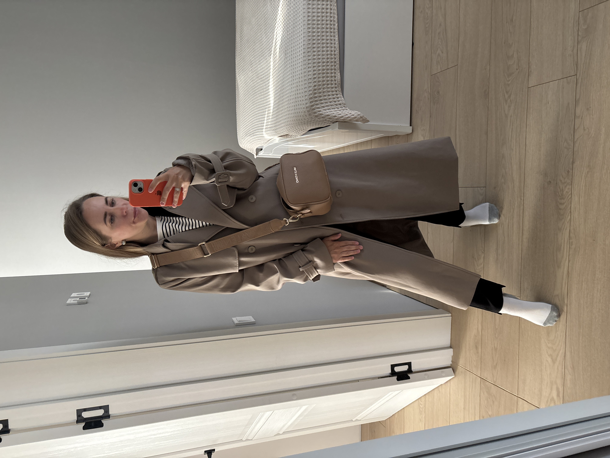 A woman is depicted taking a mirror selfie indoors. She holds an orange-cased smartphone, partially obscuring her smiling face. She has brown hair and wears a light beige trench coat, a black and white striped top, black pants, and white socks with grey toes.
A tan crossbody bag with a broad strap is worn across her body, resting on her hip. The reversed text "MOLIUIEO" is visible on the bag's flap.
The setting is a room with light grey walls and light-colored wooden floorboards. To the woman's right, part of a bed with a white, textured waffle-weave blanket is visible, illuminated by what appears to be natural light creating defined shadows. A white door with black hardware is also present in the background. The scene suggests a bright, daytime interior, possibly a residential bedroom or hallway.
