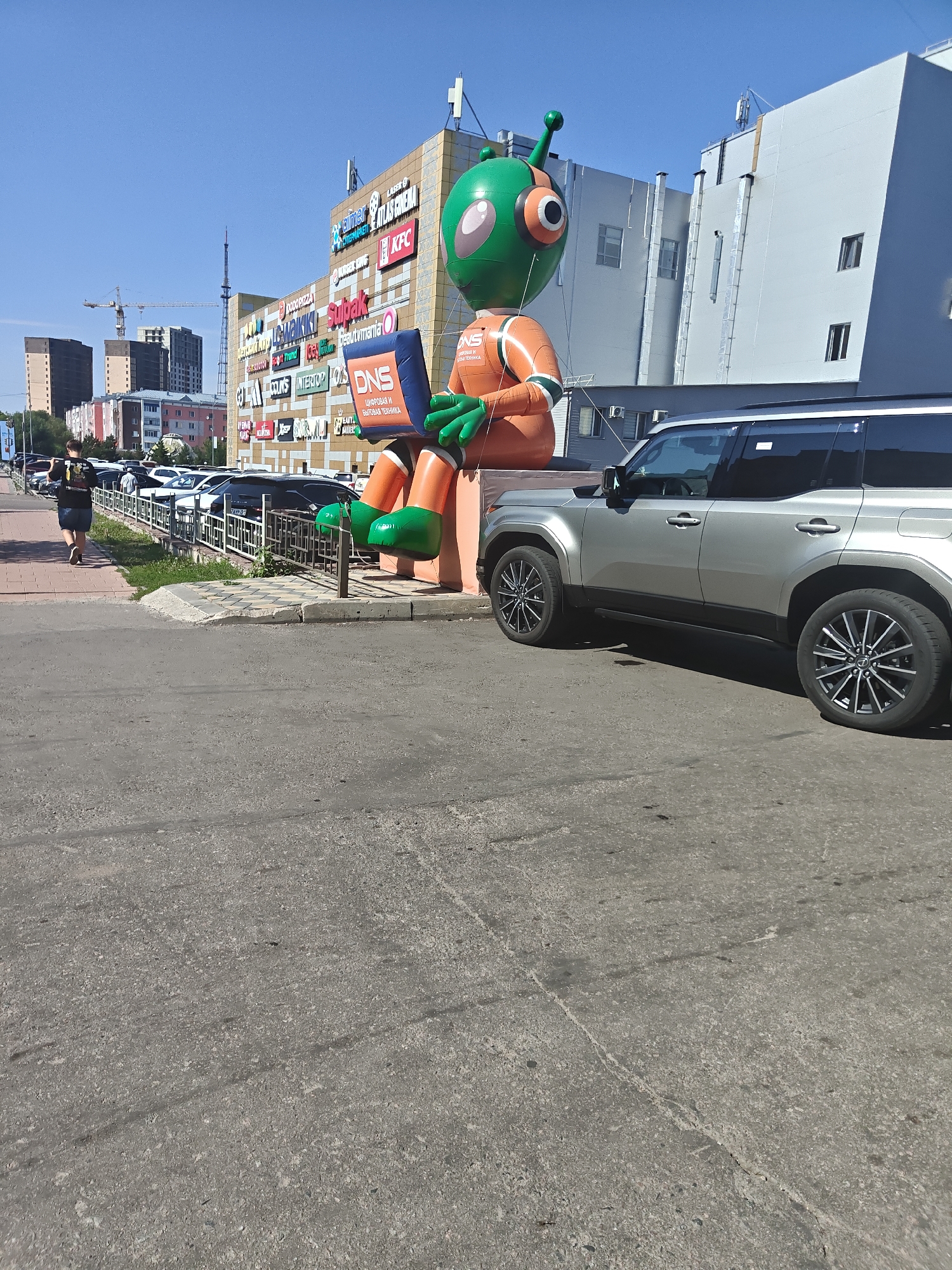 A large inflatable alien figure, primarily orange and green, stands prominently in front of a multi-story commercial building. The alien is holding a blue laptop. The building facade is adorned with numerous signage for various businesses including "DNS," "Burger King," "KFC," "Subway," and "Cinema." A silver SUV is parked to the right of the alien. In the background, a line of parked cars is visible along a street, and further in the distance, high-rise buildings and construction cranes suggest an urban environment. A person is walking away from the camera on a sidewalk to the left of the scene. The weather appears to be sunny with a clear blue sky. The overall setting is a streetscape in the city of Petropavl, Kazakhstan, likely in a commercial district.
