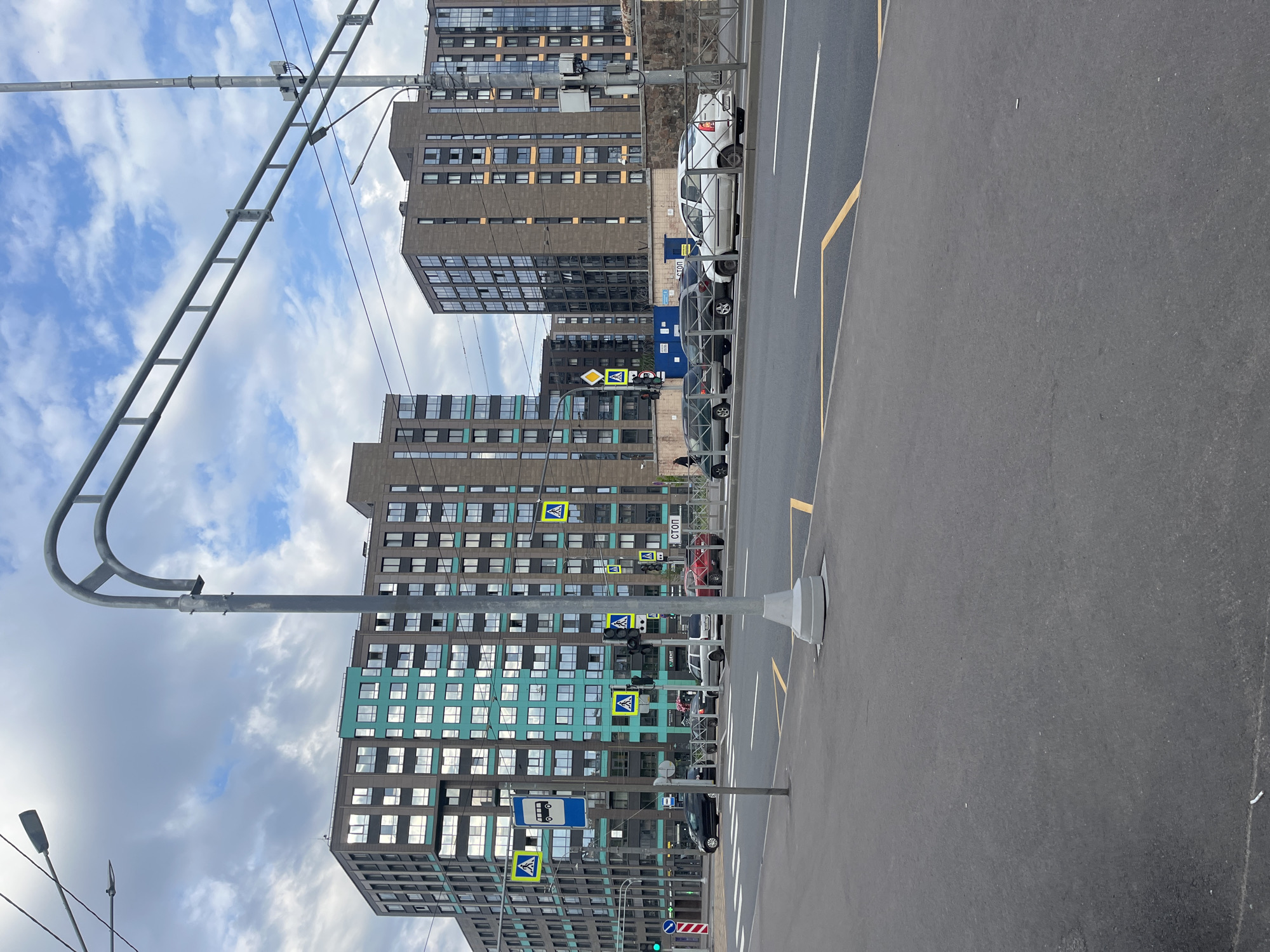 The image depicts a modern urban street scene during the daytime, under a partly cloudy sky. On the left side, several tall, multi-story buildings dominate the background. These buildings feature numerous rectangular windows and vary in facade color; some are dark brown or grey, while others, particularly one prominent building, incorporate sections of turquoise/teal paneling.
In the foreground and extending towards the right, there is an asphalt road with clear white and yellow lane markings. Several cars are parked parallel to the curb on the right side of the street, including white, black, and dark grey vehicles.
The street is equipped with typical urban infrastructure. A large metal gantry-like structure spans across the top of the frame, possibly for street lighting or traffic signal support. Multiple streetlights and traffic signals are visible. Several yellow diamond-shaped signs with a black pedestrian symbol, indicating pedestrian crossings, are prominent. A blue rectangular sign with a white bus icon marks a bus stop. One white rectangular sign with a red border clearly displays the word "СТОП" (STOP) in Cyrillic letters. Another sign, partially obscured, seems to have "СТРОЙ" visible. Overhead wires crisscross the sky, suggesting potential electric transport lines or utility cables. No people are visibly present, and the scene appears calm and functional.