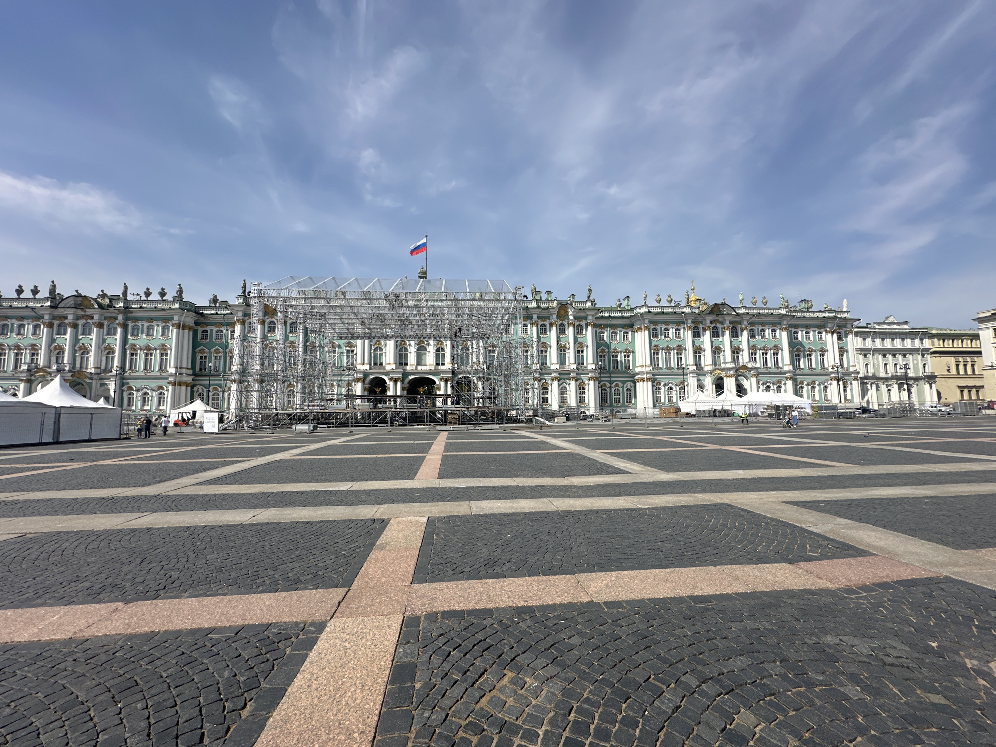 The image captures the Winter Palace in Saint Petersburg, Russia, viewed from Palace Square. The palace's facade is primarily green and white, with scaffolding covering a portion of the building. The Russian flag flies atop the scaffolding. The square's paving is patterned with alternating dark and light stones. Several white tents are set up in the square. A few people are visible in the square. The sky is blue with scattered clouds.